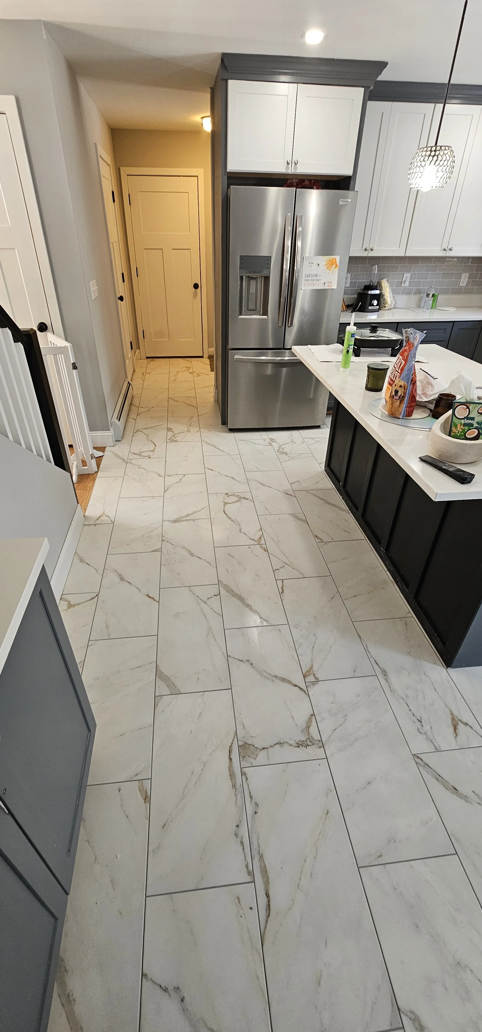Kitchen with grey and white cabinets, stainless steel refrigerator, marble-like tile floor, kitchen countertop and scattered items.