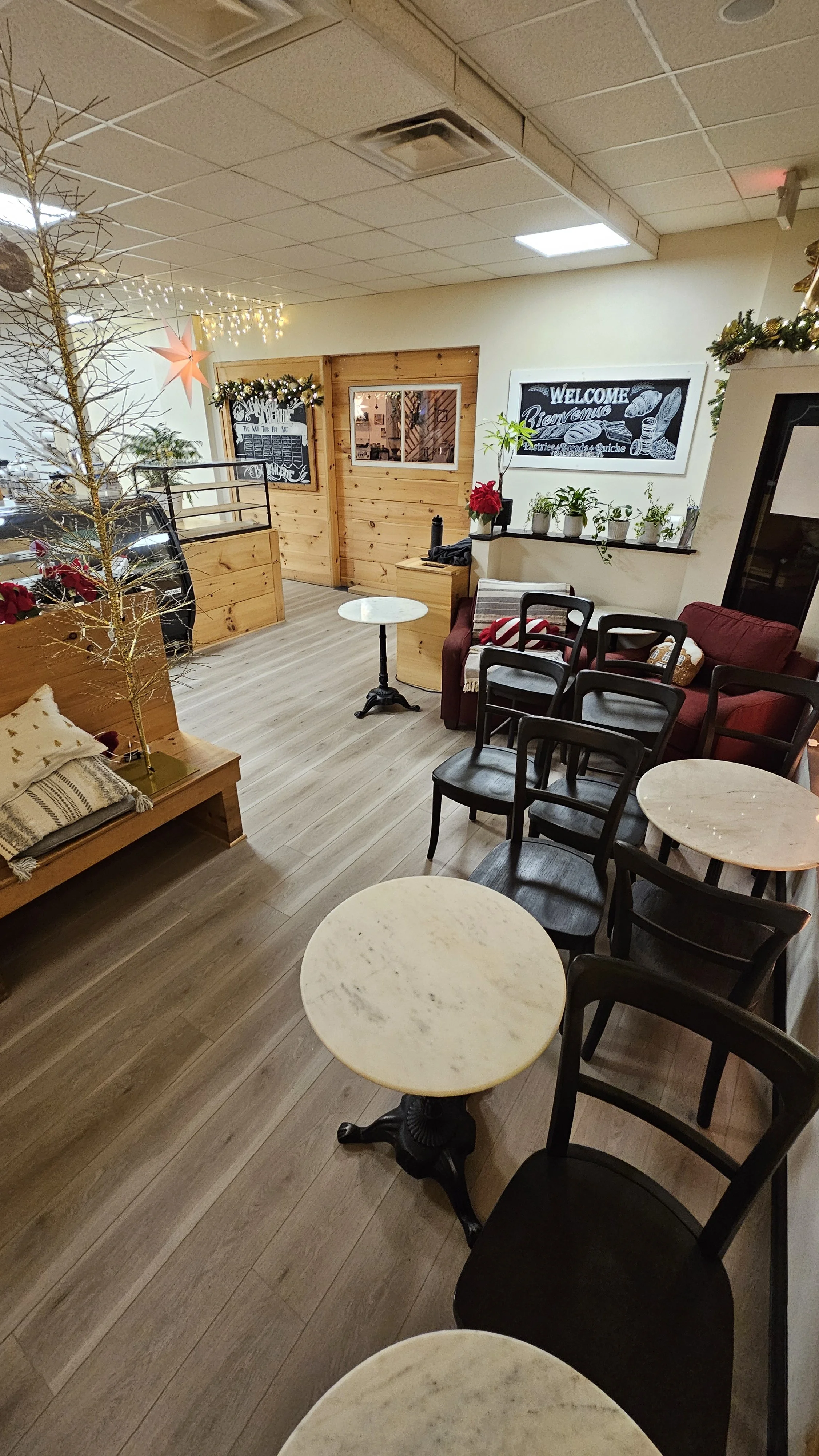 A cozy decorated indoor space with black, white, and red furniture, small round tables, potted plants, and Christmas decorations including a gold tree, stars, and garlands.