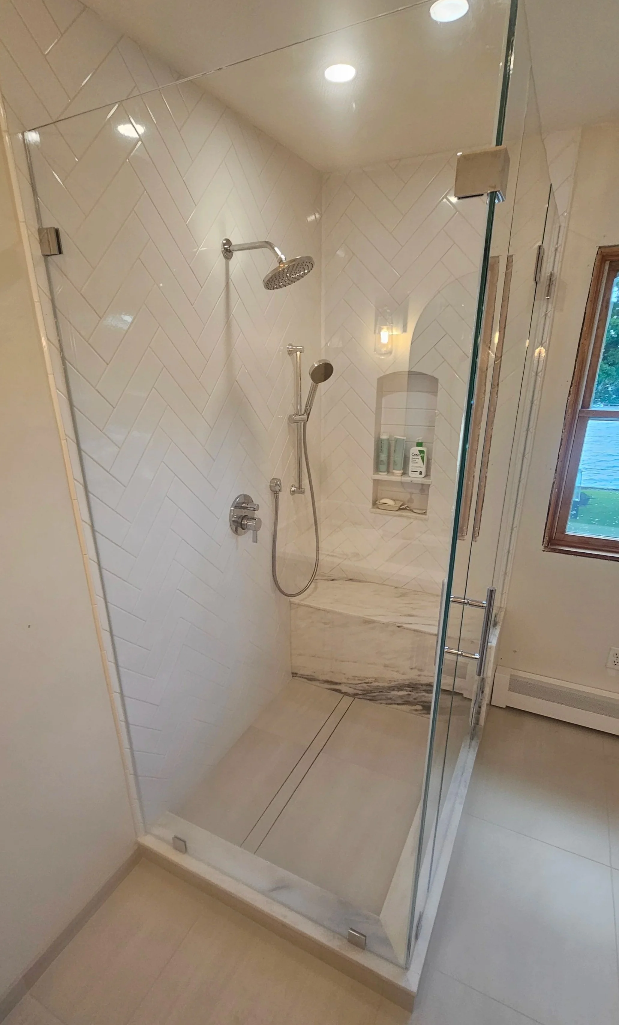 A modern shower with white herringbone tile walls, a rain showerhead, a handheld shower, and a glass door. There is a built-in shower shelf with toiletries and a window with a view outside.