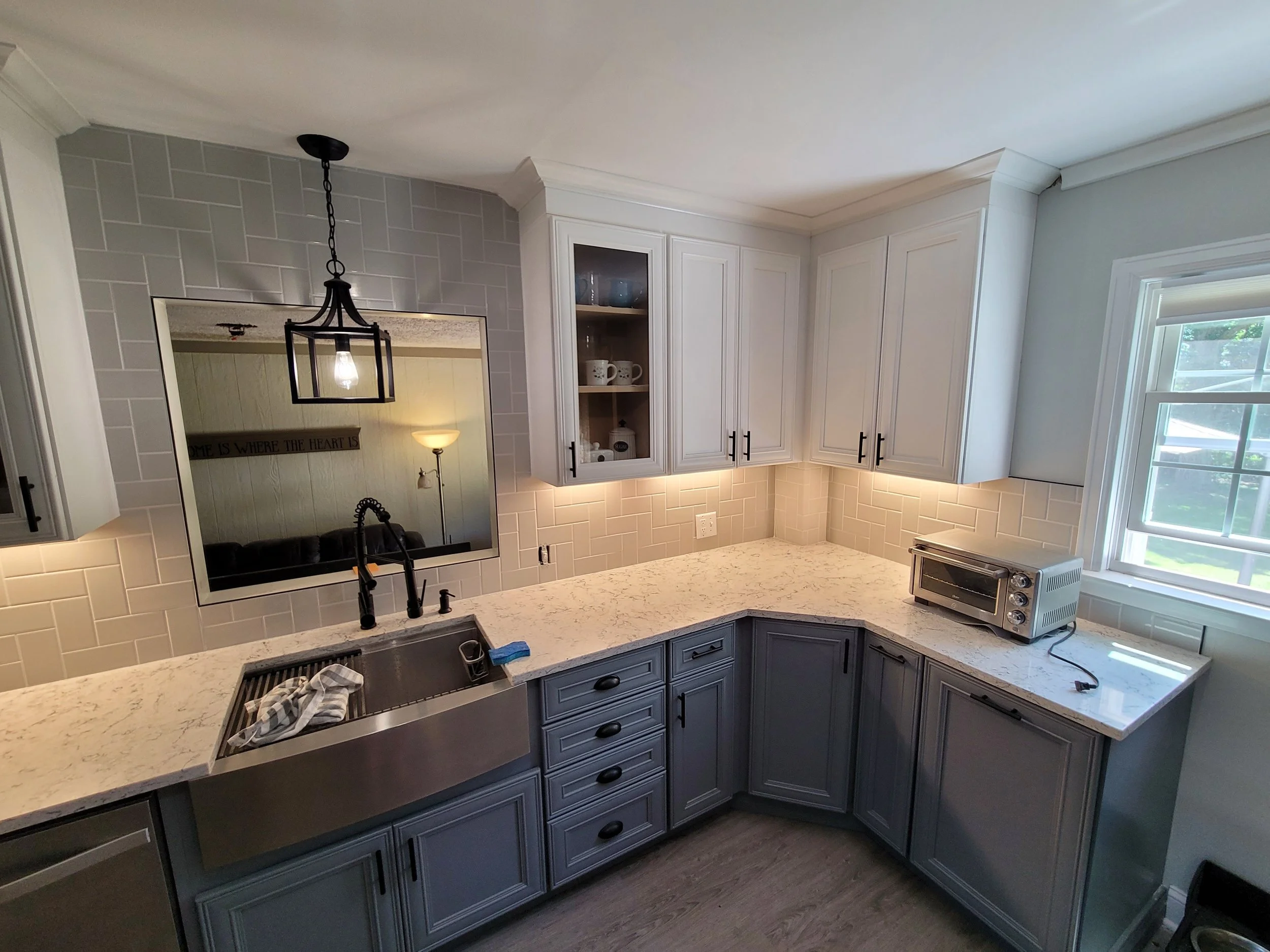 A kitchen with white cabinets, light-colored granite countertops, and a window letting in natural light. There is a small toaster oven on the counter, a black sink faucet, and a mirror on the wall reflecting part of the room.