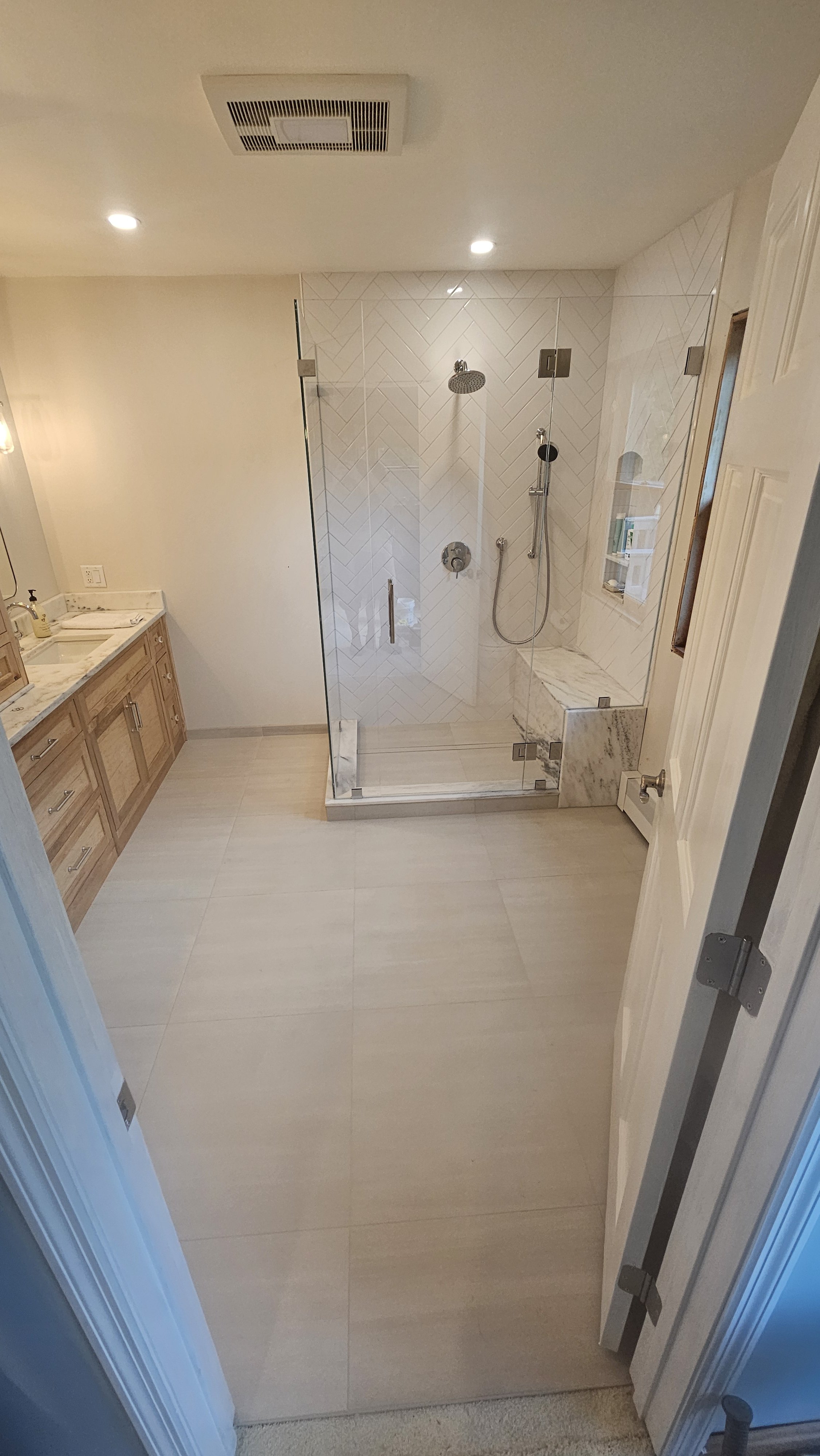 A modern bathroom with a glass-enclosed shower, wooden vanity with a marble countertop, and beige floor and wall tiles.