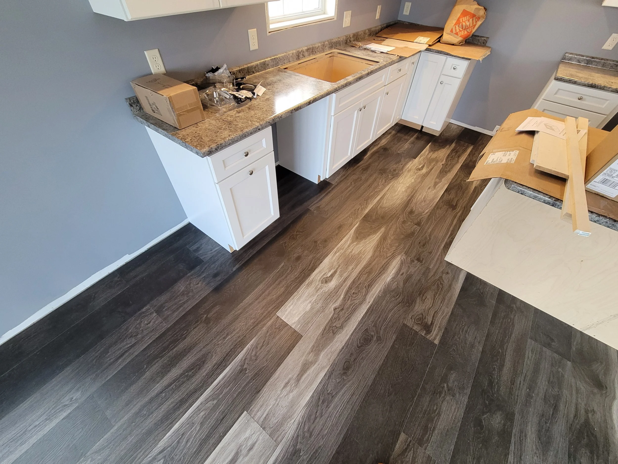 Kitchen under renovation with new dark wood flooring, white cabinets, and a granite countertop. Packaging and tools are scattered on the counters.