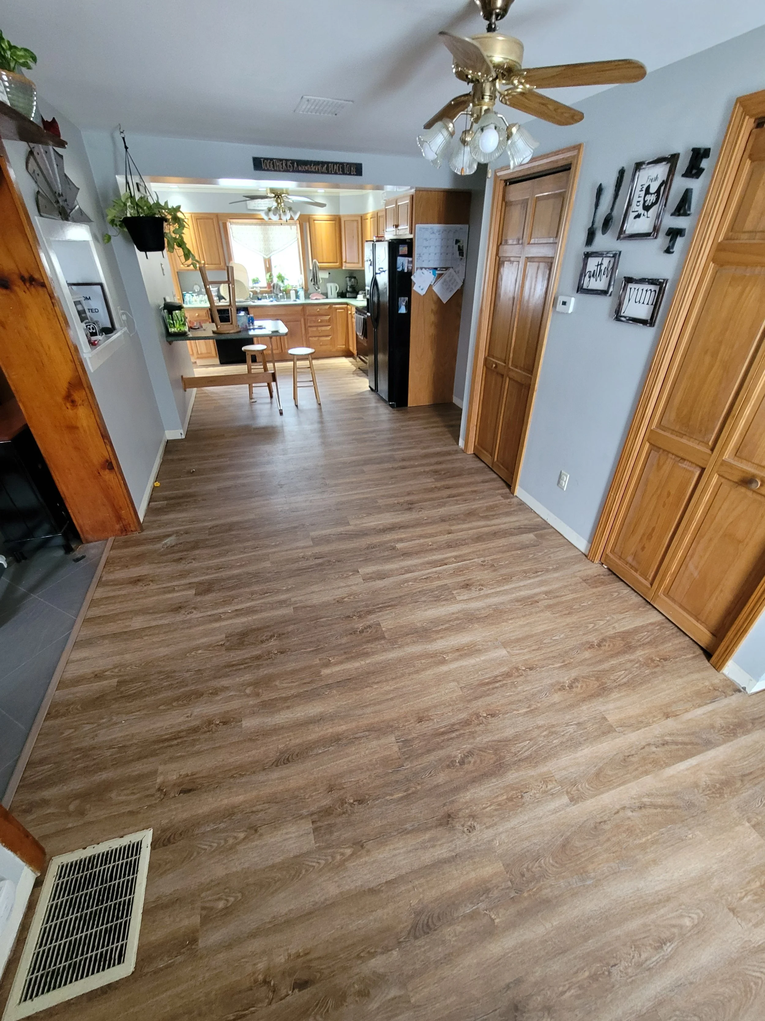A view of a kitchen and dining area with wood flooring, wooden cabinets, a refrigerator, and a small counter with stools. Decor includes wall art with words like "Laugh," "Grateful," and "Yum" and household items such as a hanging plant, a kitchen wi