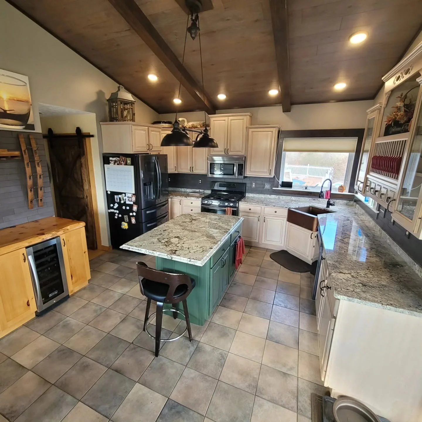 A spacious kitchen with a wooden ceiling, recessed lighting, granite countertops, and a central island with a green base. It features white cabinets, a black refrigerator, microwave, and stove. There is a window above the sink, a small wine fridge, a