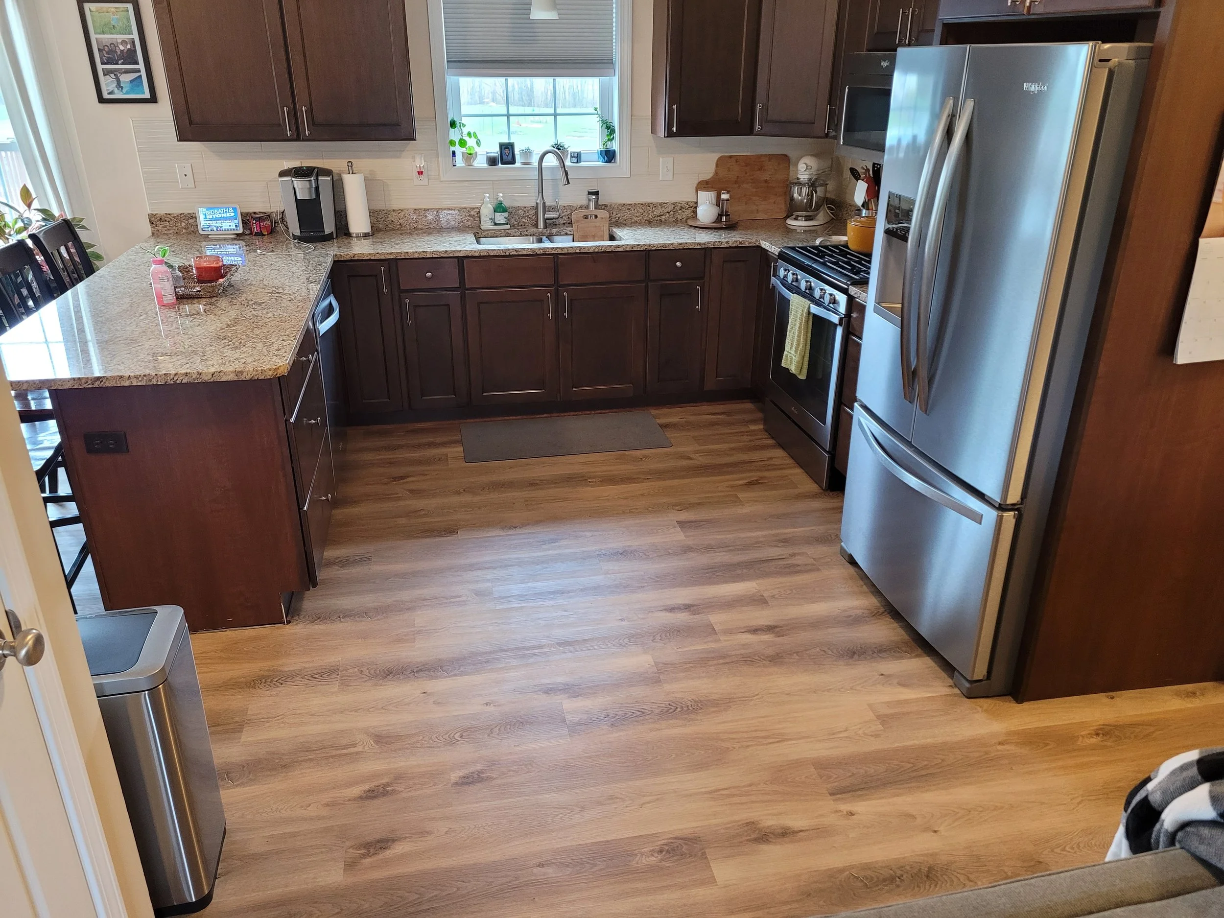 A kitchen with dark wooden cabinets, granite countertops, a window above the sink with plants and framed photos, stainless steel appliances including a refrigerator, stove, and microwave, and various kitchen items on the counter.