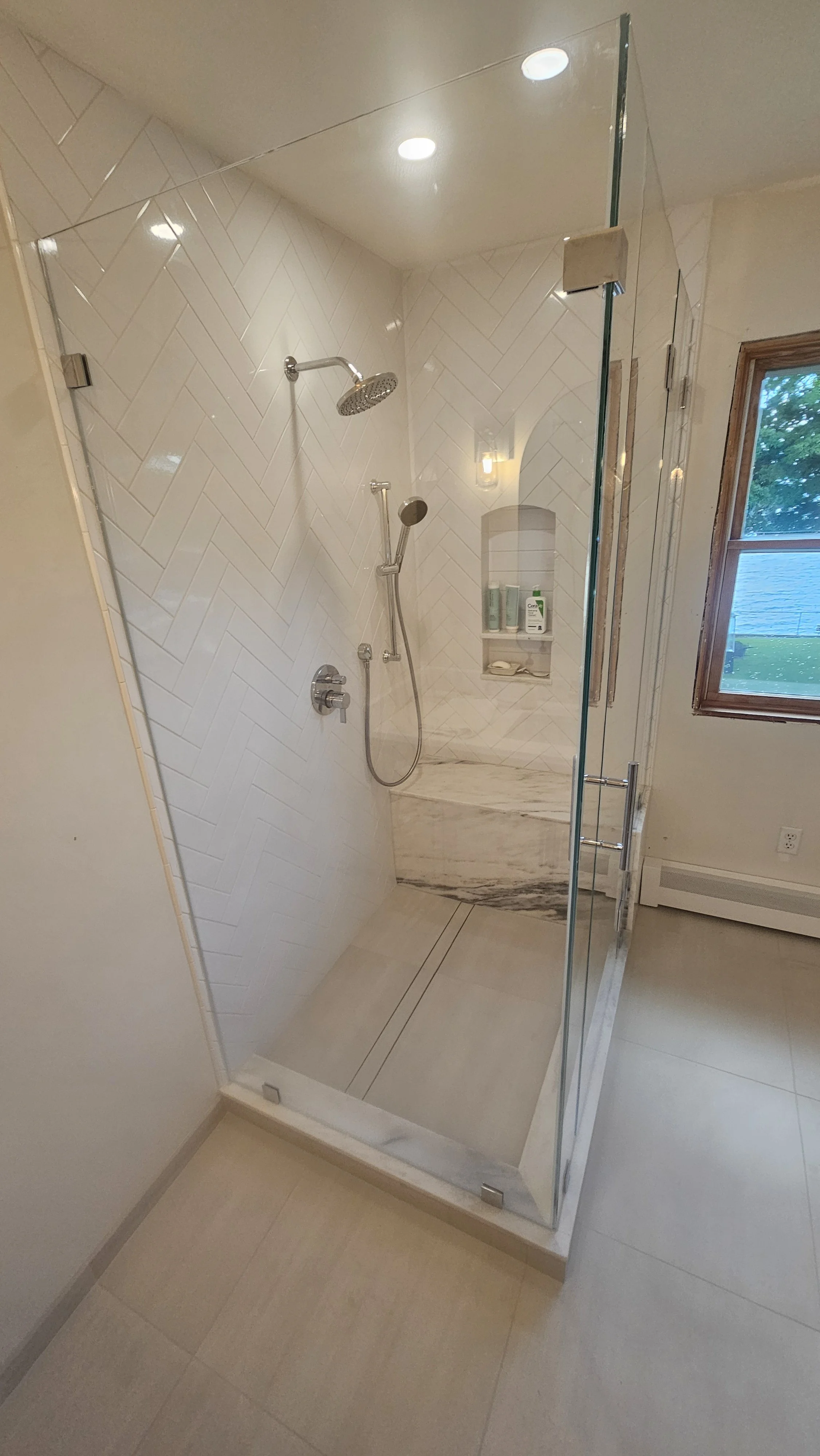 A modern walk-in shower with white herringbone tile on the wall, a rainfall showerhead, a handheld shower, a built-in niche with toiletries, and a glass door, with a window showing greenery outside.