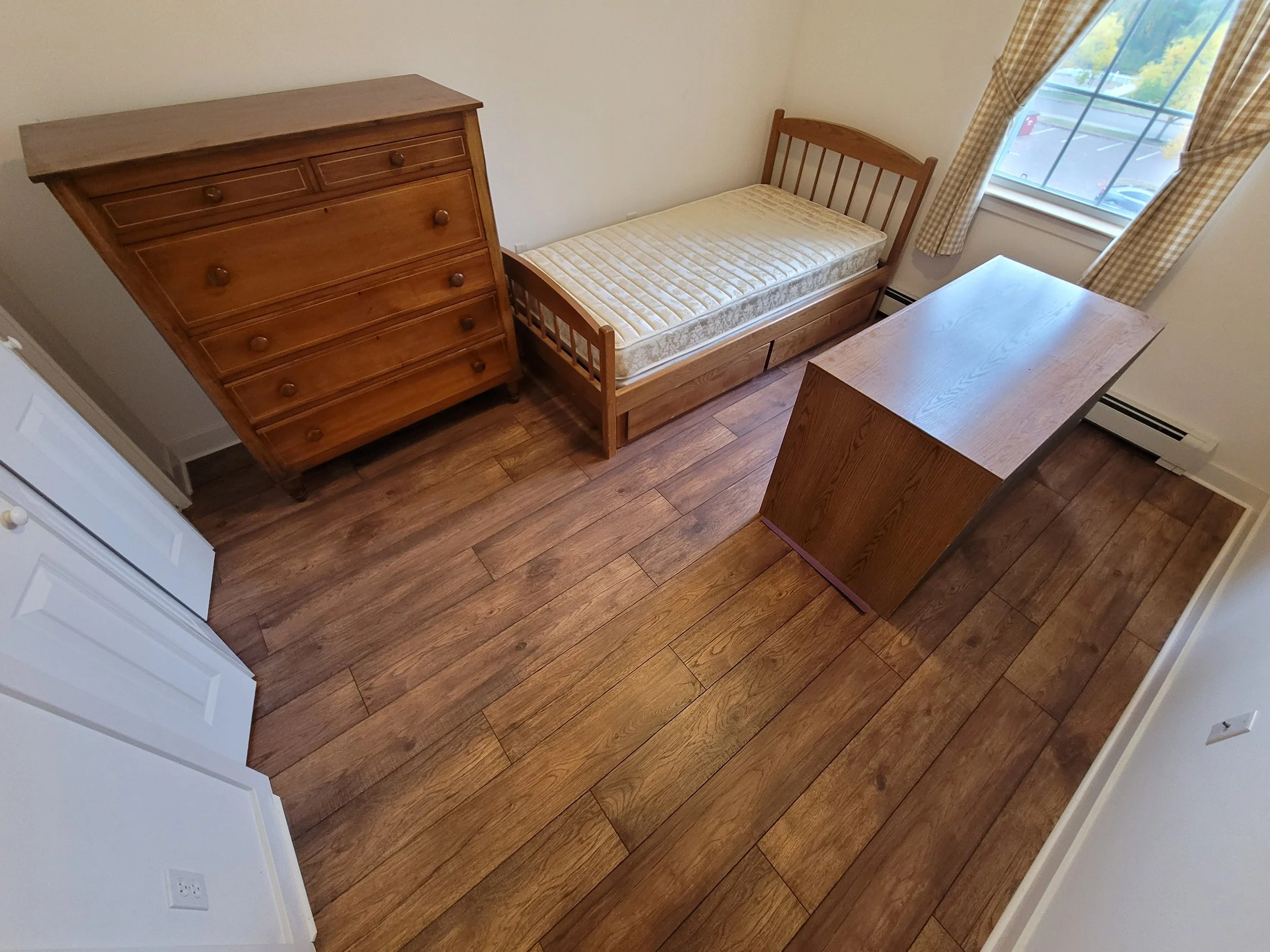 Empty bedroom with hardwood floors, a wooden dresser, a bed with an unmade mattress, a desk, a window with checkered curtains, and a heater along the wall.