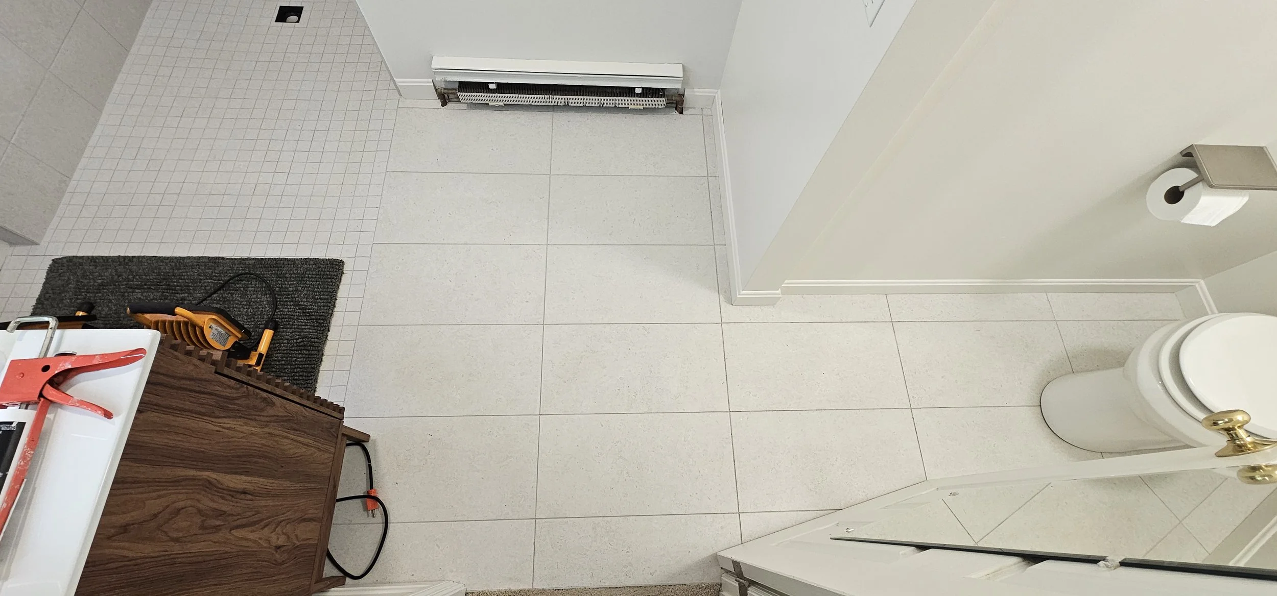 View of a bathroom floor with white tiles, a small white tiled section near the door, a black floor mat, and a wooden cabinet with tools on top. Part of a toilet and a toilet paper roll on the wall are visible.