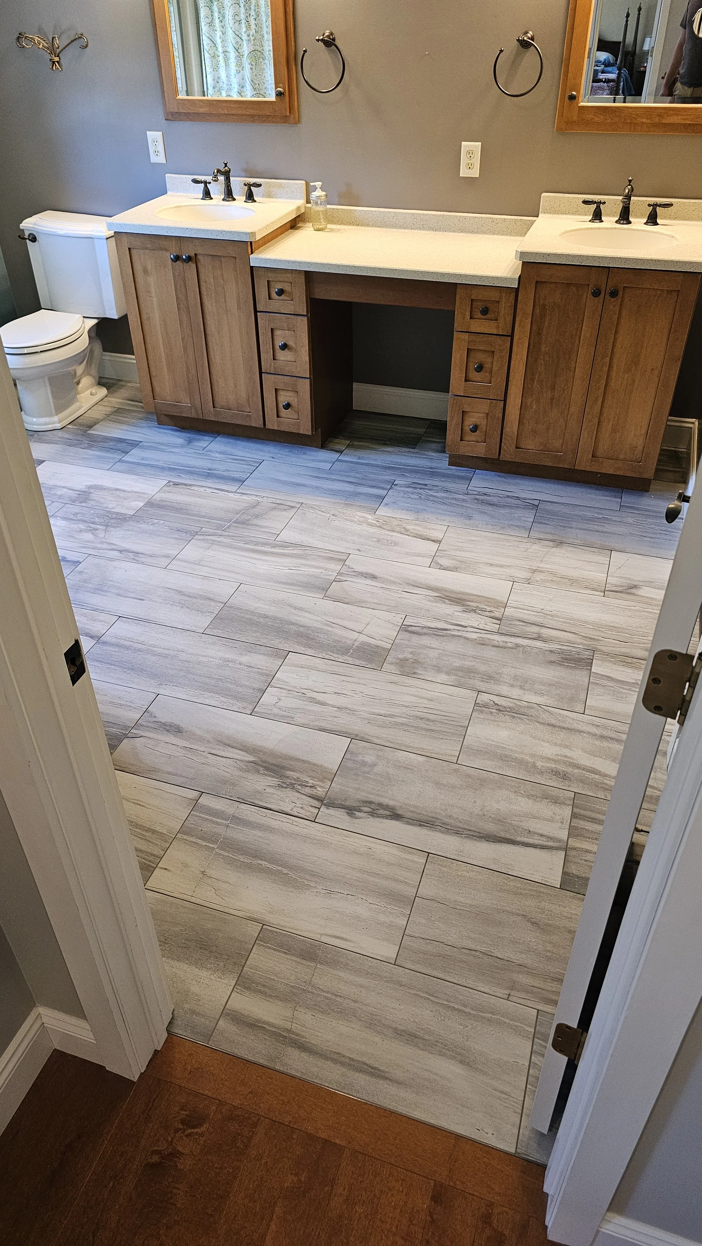 Bathroom with gray walls, two wooden vanities with white countertops and black fixtures, three drawers each, one needs a sink, two mirrors with wooden frames, two towel rings, and tiled floor with gray and white patterns.