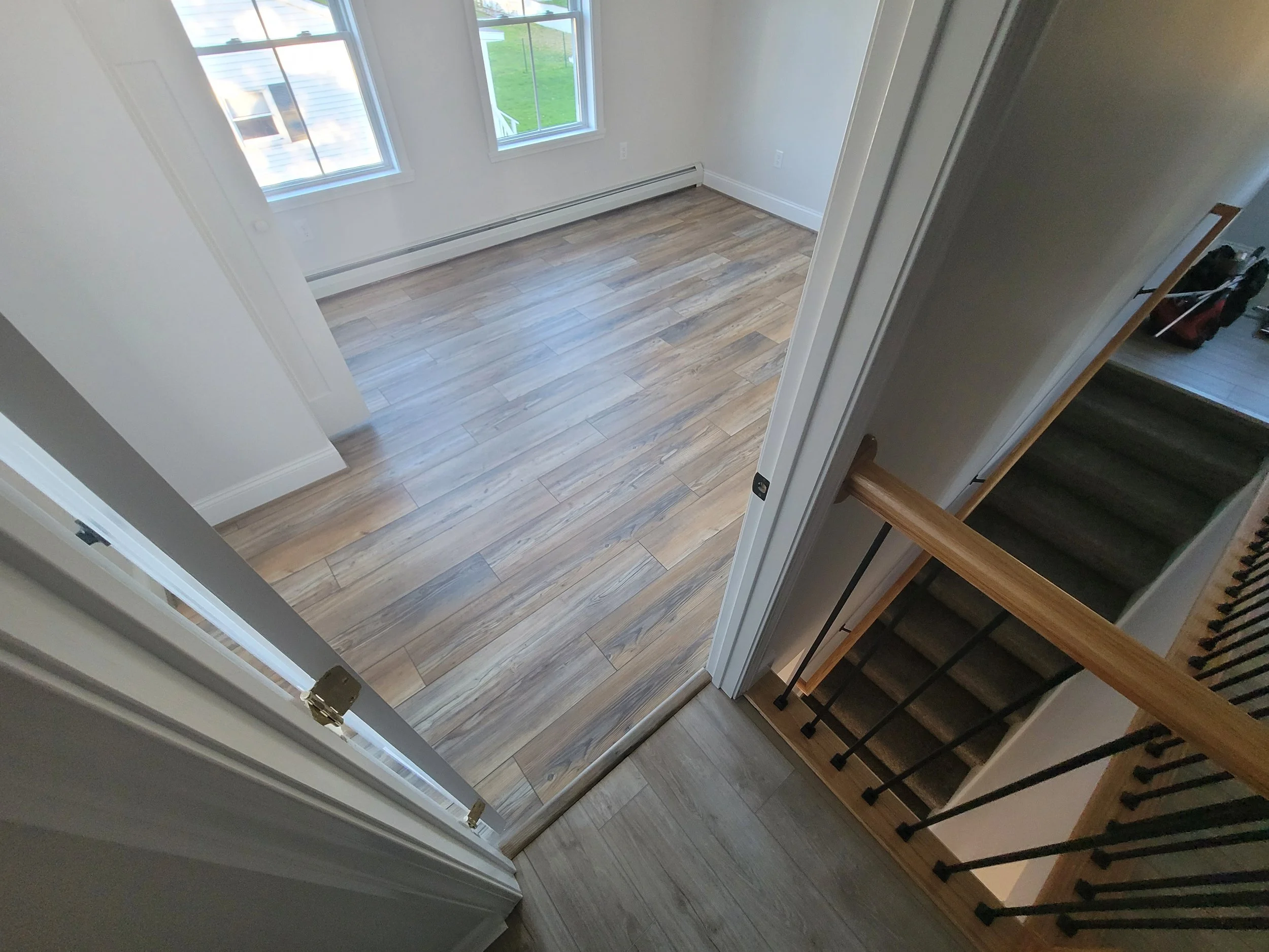 View from the top of a staircase looking into a room with wooden flooring, white walls, and two windows showing green outdoor space.