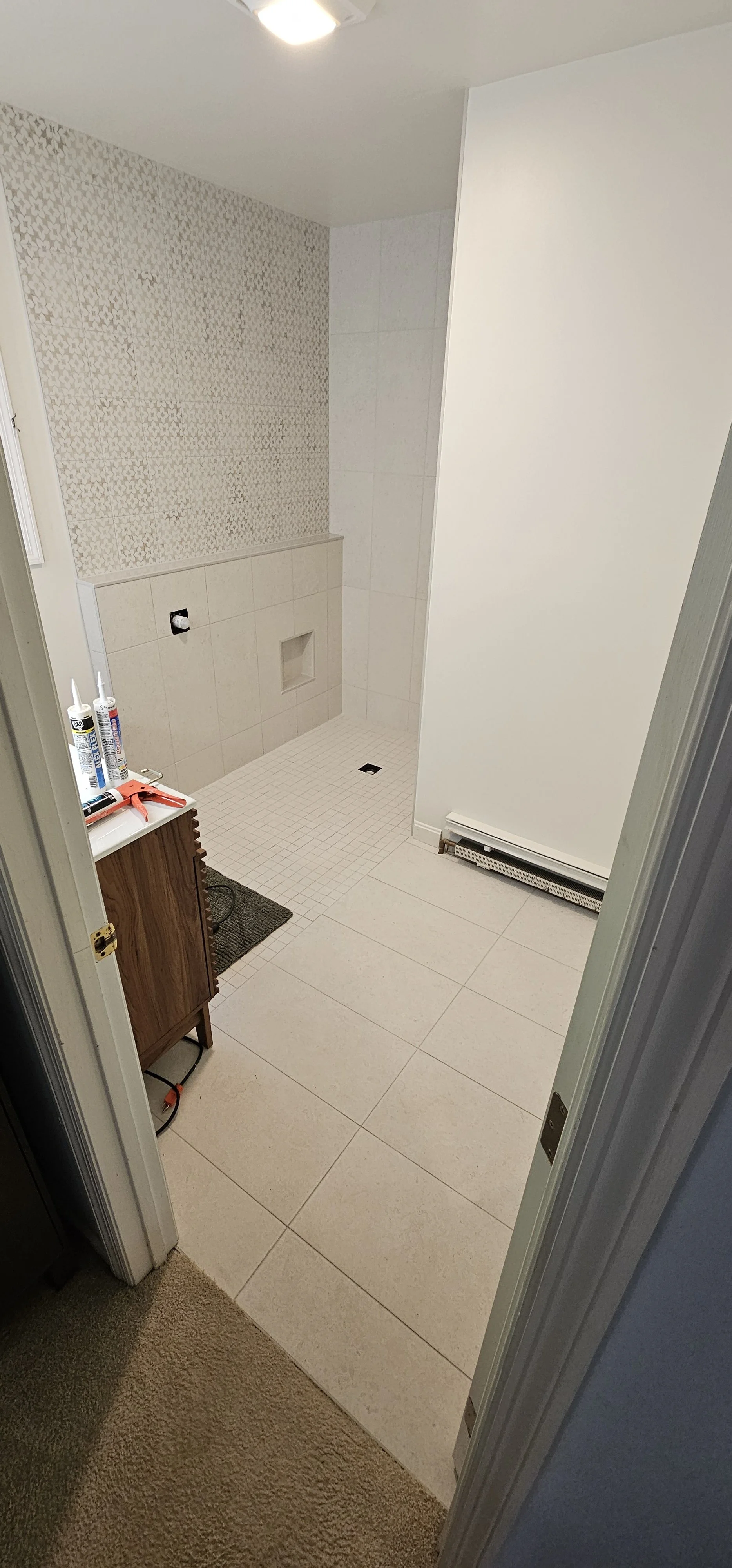 Bathroom under construction with tiled shower area, no fixtures installed, construction supplies on a small wooden cabinet, and a baseboard heater.