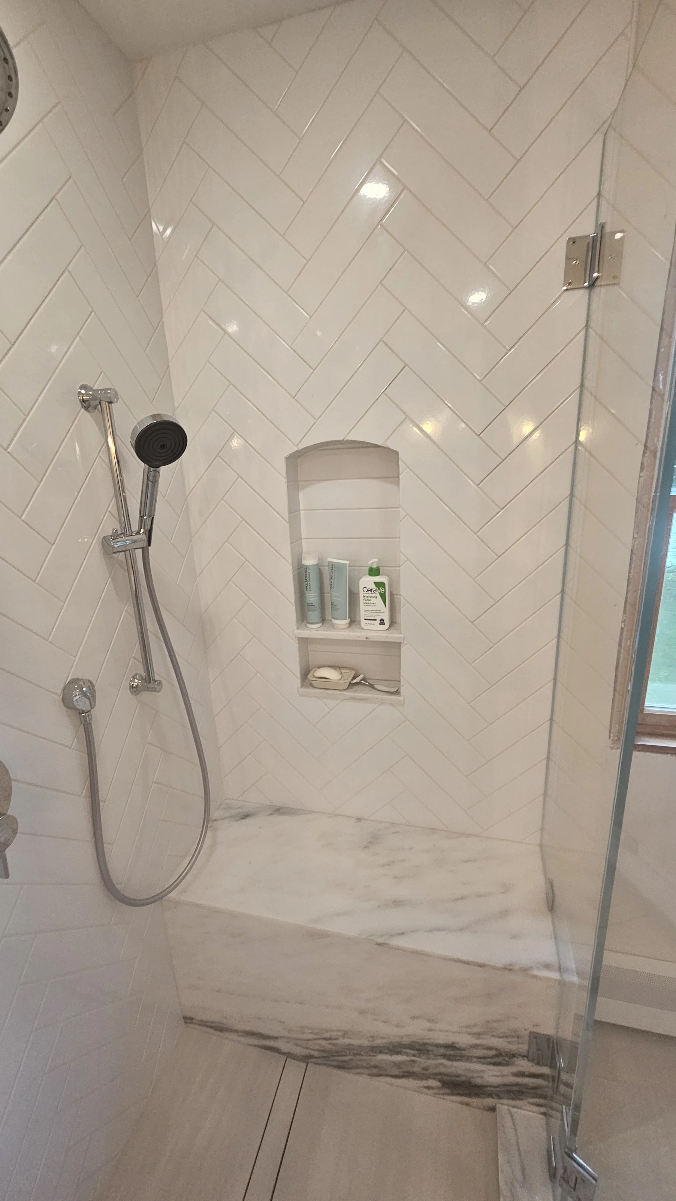 Shower area with herringbone tiled wall, a marble shelf with toiletries, a handheld shower head, and a glass shower door.