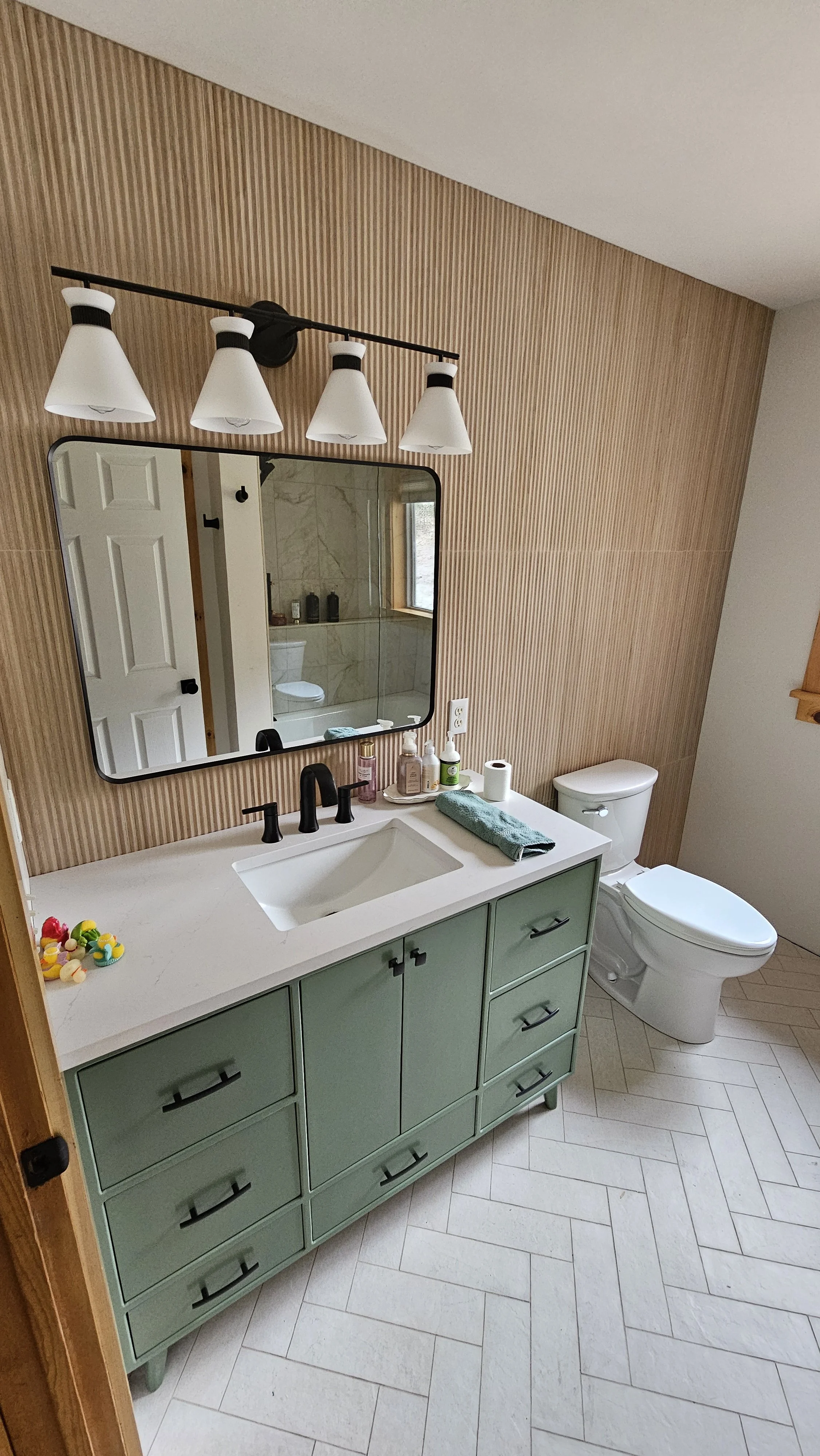 Bathroom with a light green vanity, white countertop, and a large wall mirror. The wall behind the mirror has light wooden paneling. There are four light fixtures above the mirror. The bathroom has a window, a white toilet, and various toiletries and