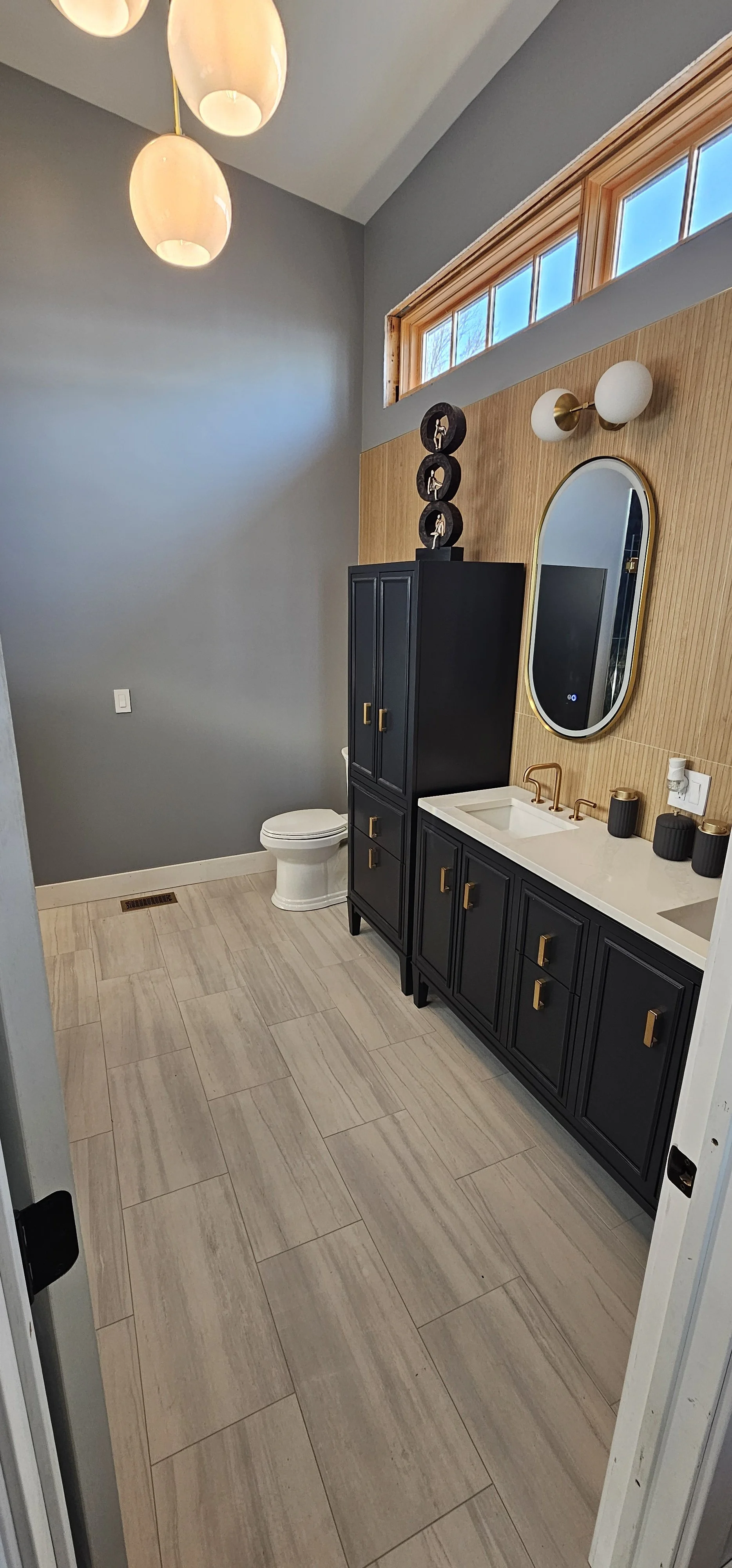 A modern bathroom with a gray wall, beige tile flooring, a white toilet, a black vanity with gold handles and two sinks, a large mirror, a black cabinet, and decorative black jars on the countertop. There's a high window with wooden trim and a high-m