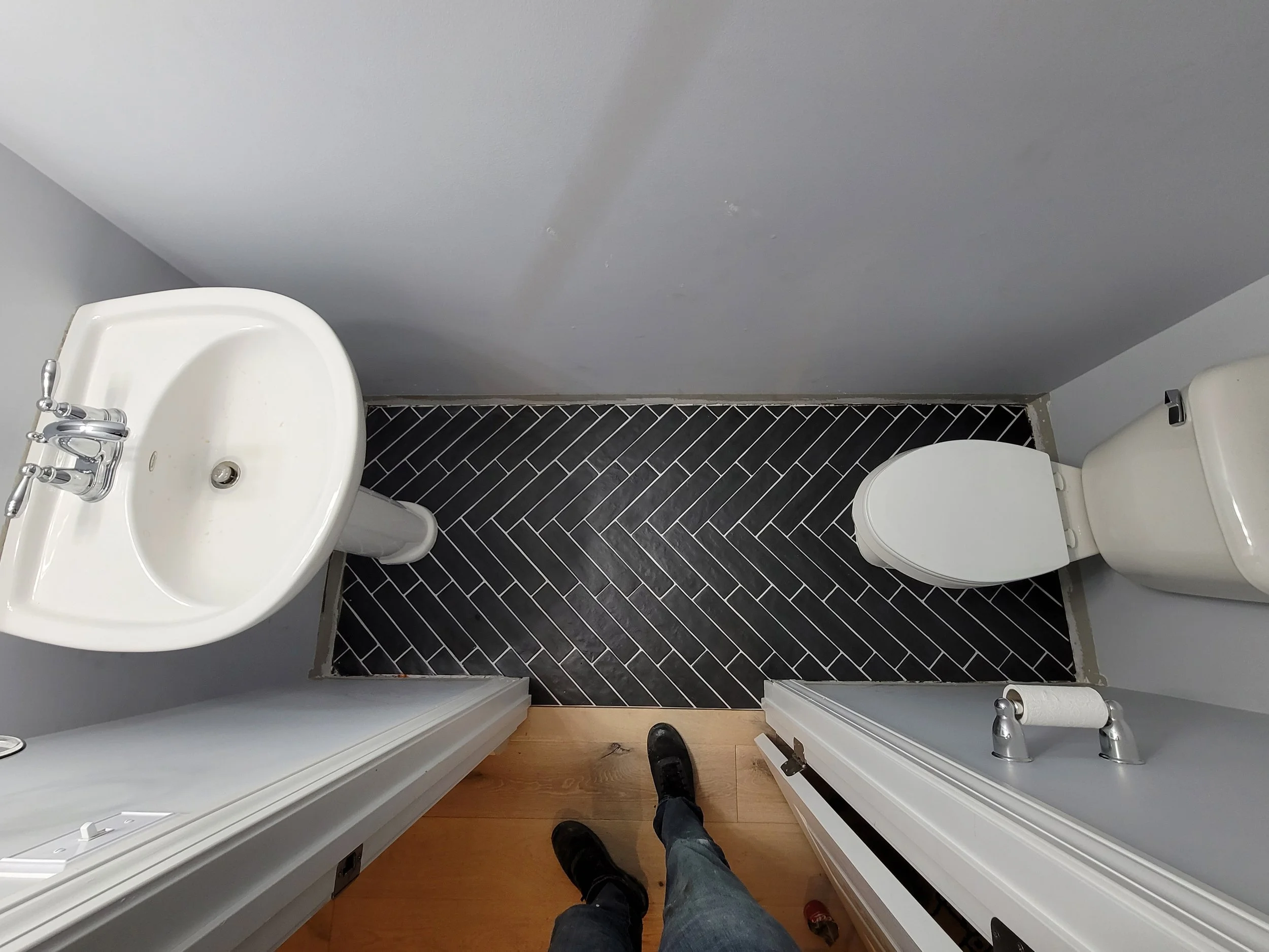 First-person perspective of a person standing inside a small bathroom or closet, with a partial view of a urinal on the left, a toilet on the right, black subway tile flooring, and gray painted walls.