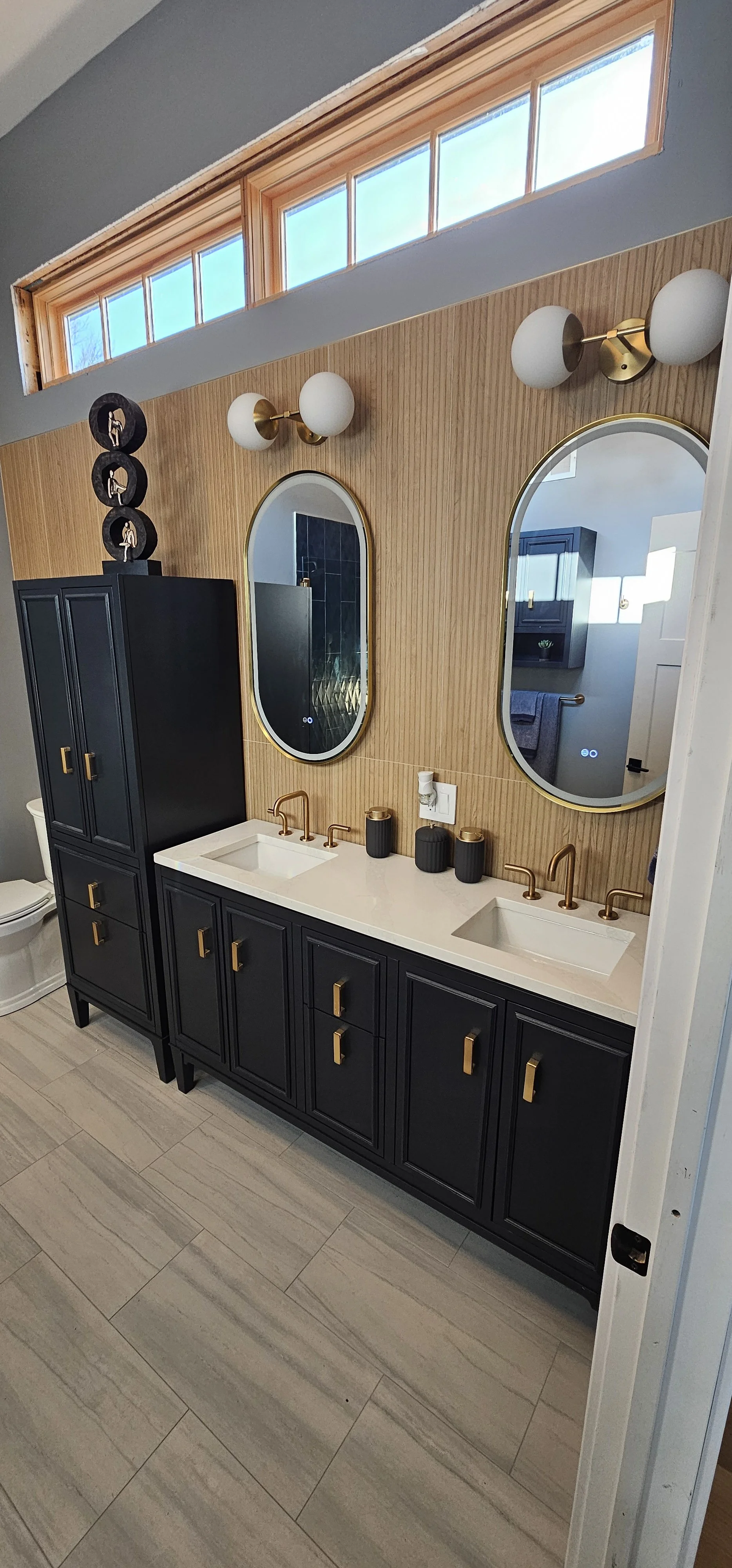 A modern bathroom with a double vanity, black cabinetry with gold handles, two oval mirrors with gold frames, bronze faucets, white countertops, and a wooden tiled wall. Above are two round sconce lights, and a window above provides natural light.