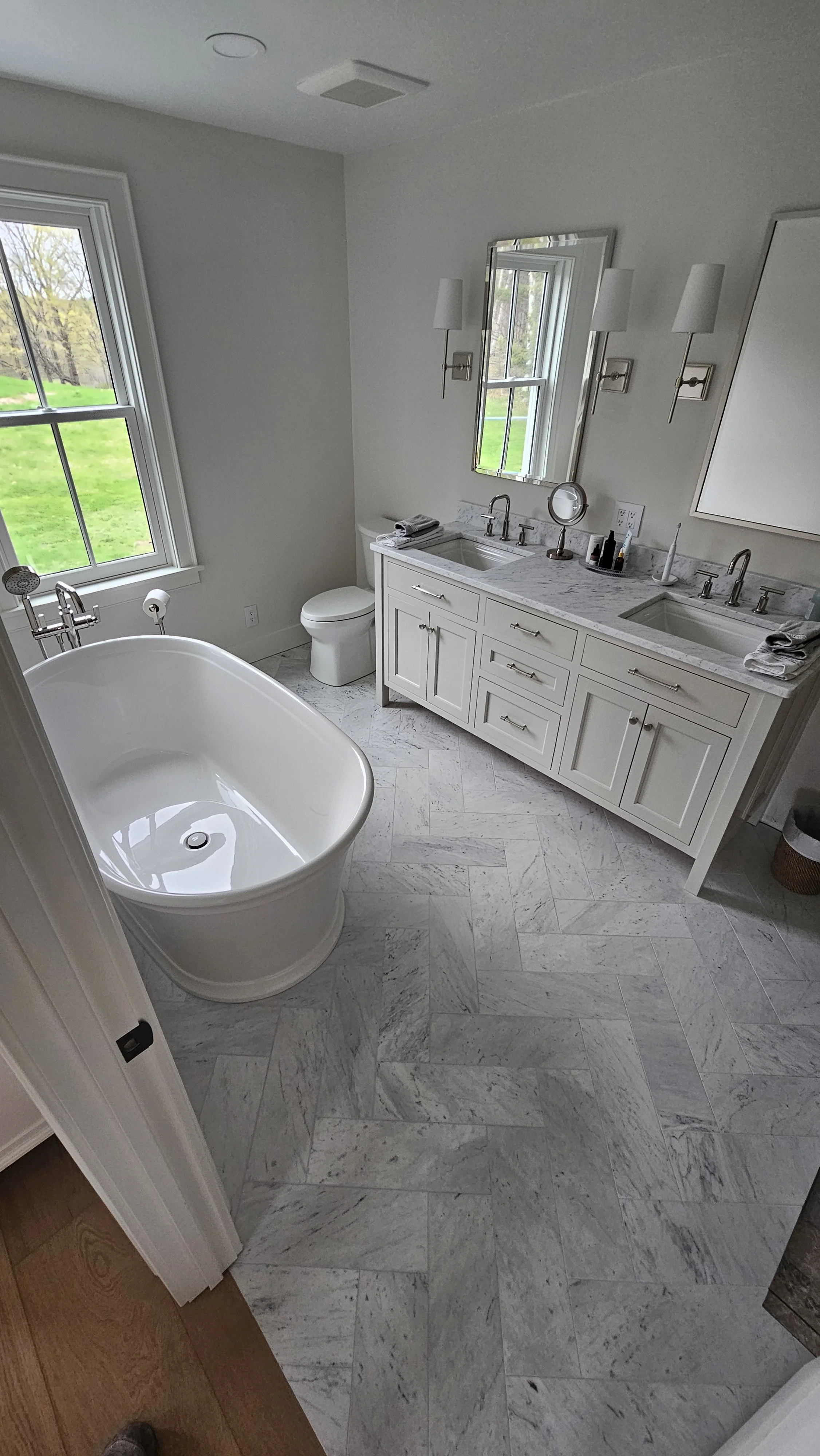 A bathroom with a white bathtub, a white toilet, a double sink vanity with a marble top, a mirror, two wall sconces, and a window showing outdoor greenery.