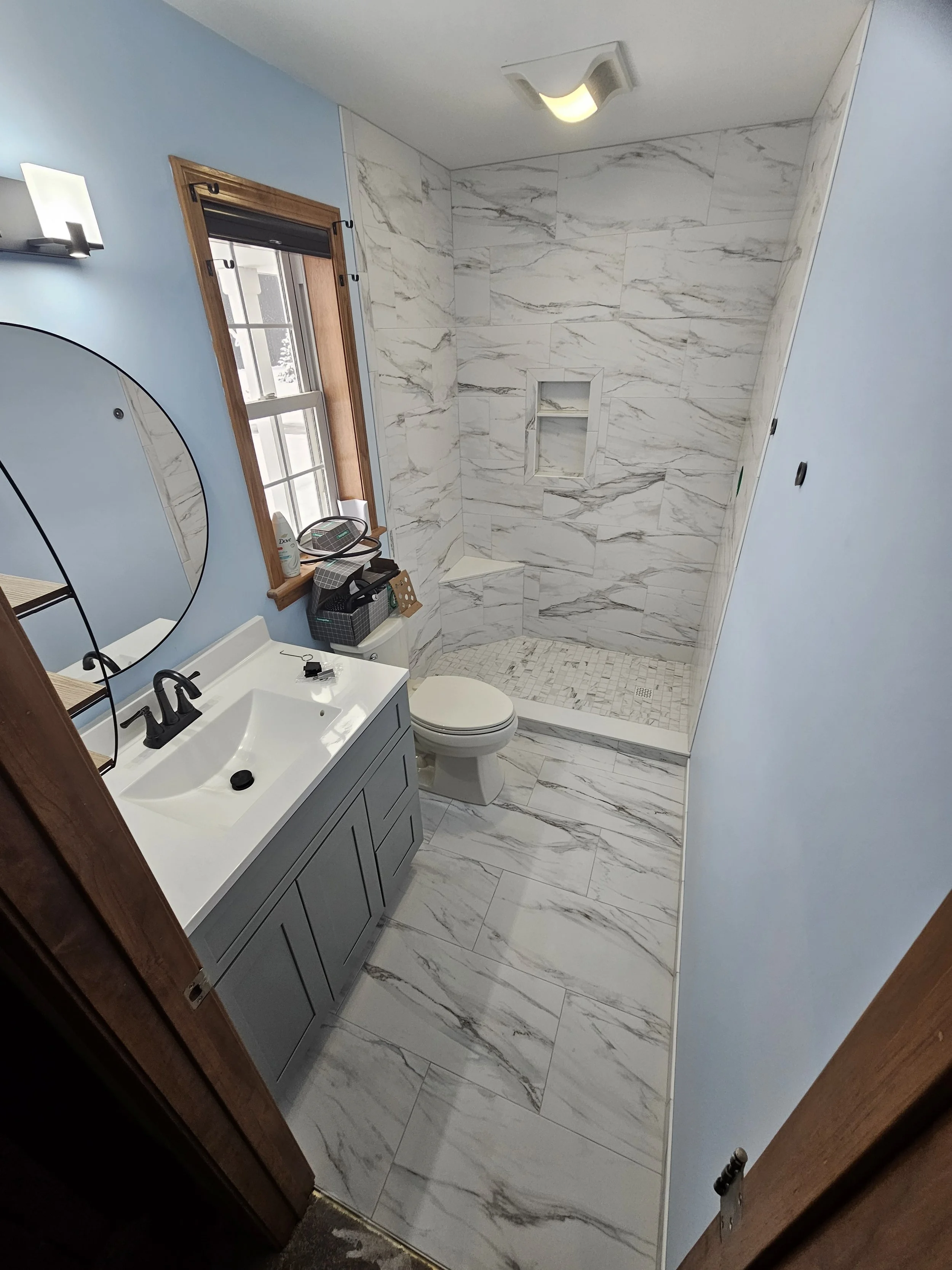 A bathroom with a light blue wall, a white sink with a black faucet, a round mirror, a window with a wooden frame, a toilet, and a marble-tiled shower area with a built-in shelf.