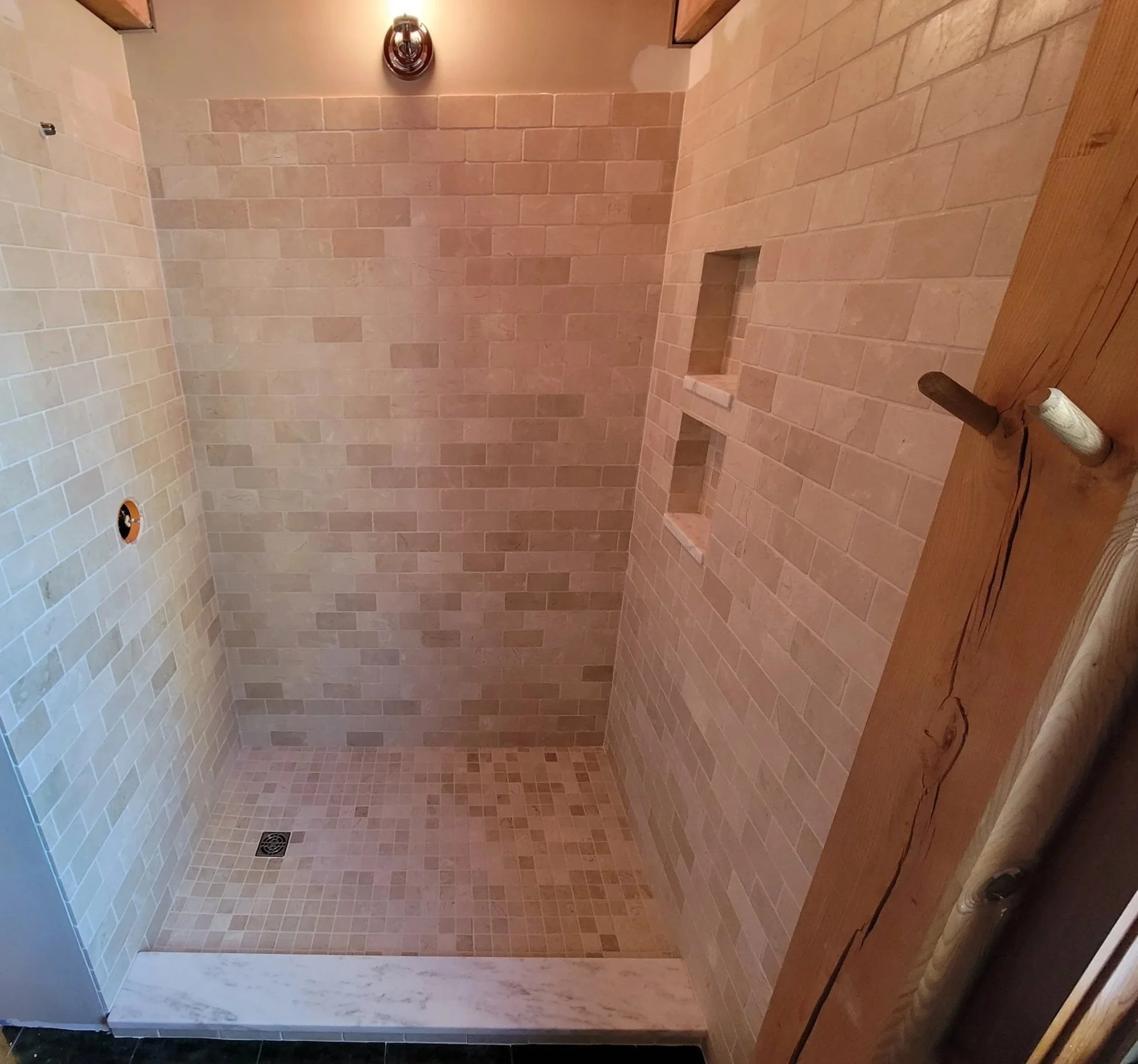 A tiled shower with beige tiles on the walls and a darker grout, two built-in shelves, a black drain on the floor, and a round ceiling drain. A wooden beam or counter edge is visible at the right, with a tool and a cord resting on it.