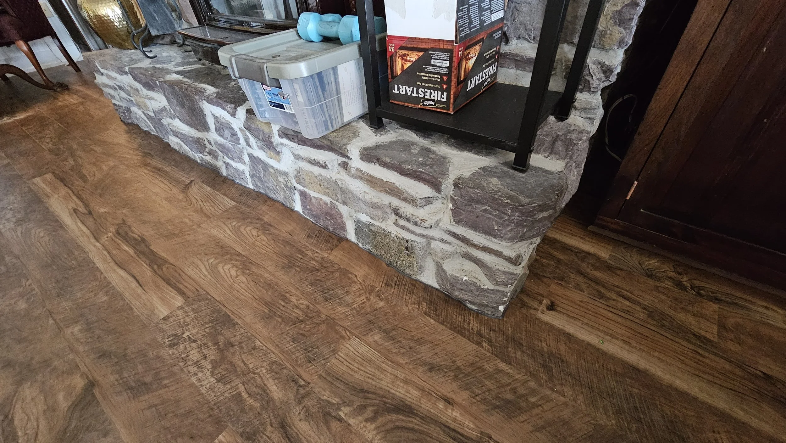 A corner fireplace with a stone hearth, surrounded by wooden flooring, with a small plastic container holding two blue dumbbells placed on it.