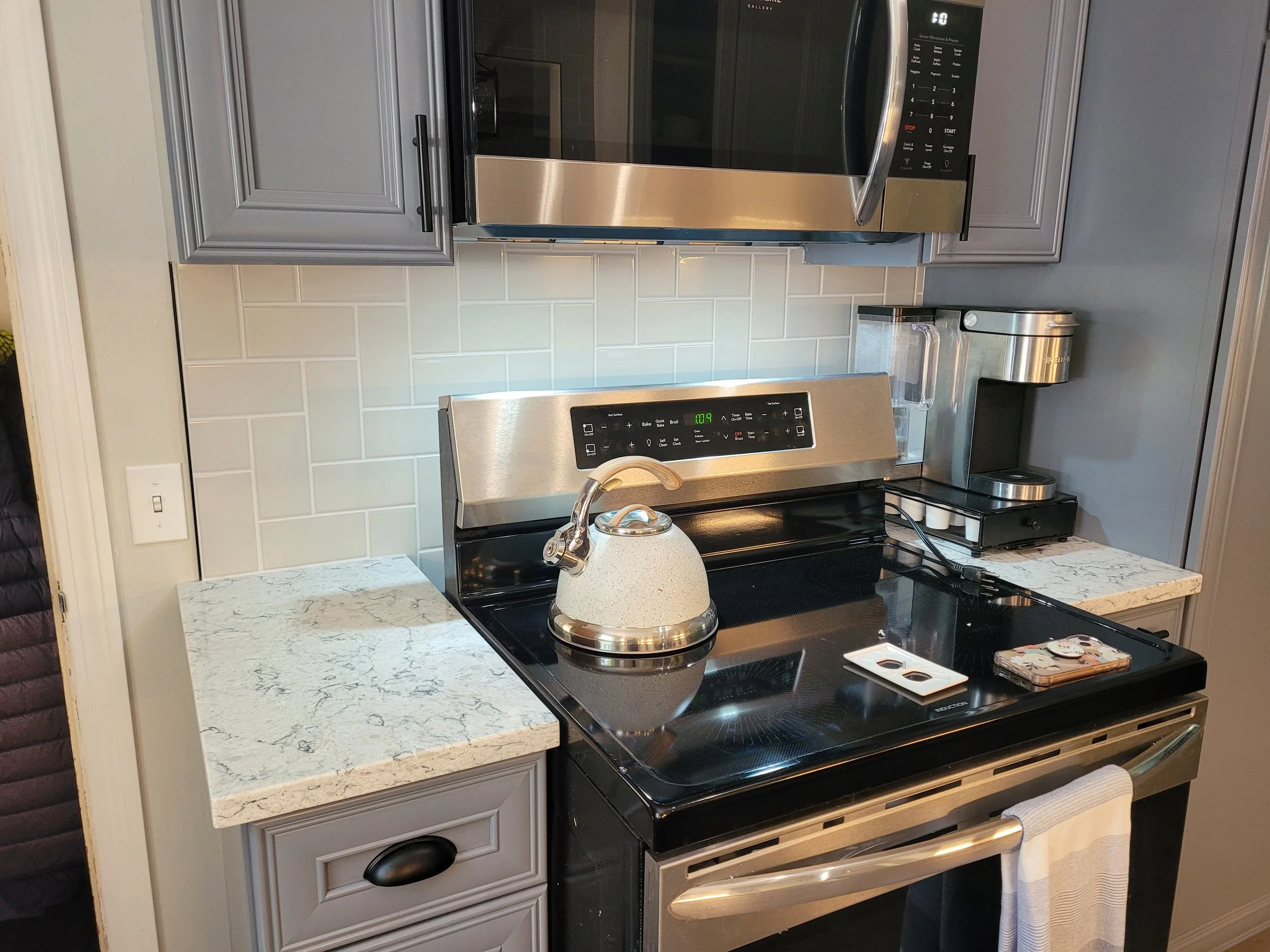 Kitchen with stainless steel microwave, stove, and kettle, gray cabinets, white tile backsplash, and granite countertop.