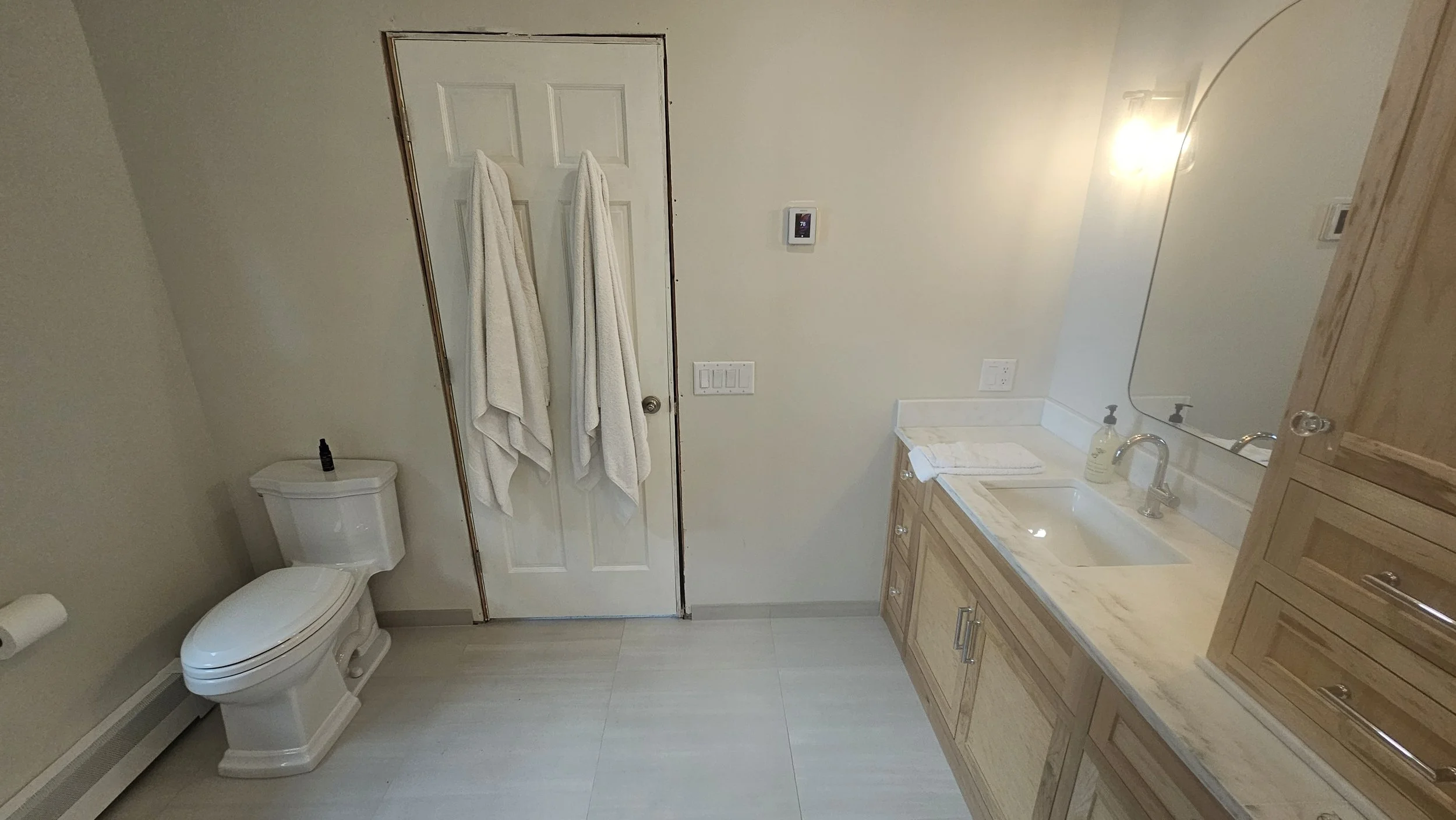 A bathroom featuring a toilet with a small bottle on top, two white towels hanging on the back of a door, a digital thermostat on the wall, a marble countertop sink, a mirror, and wooden cabinets.
