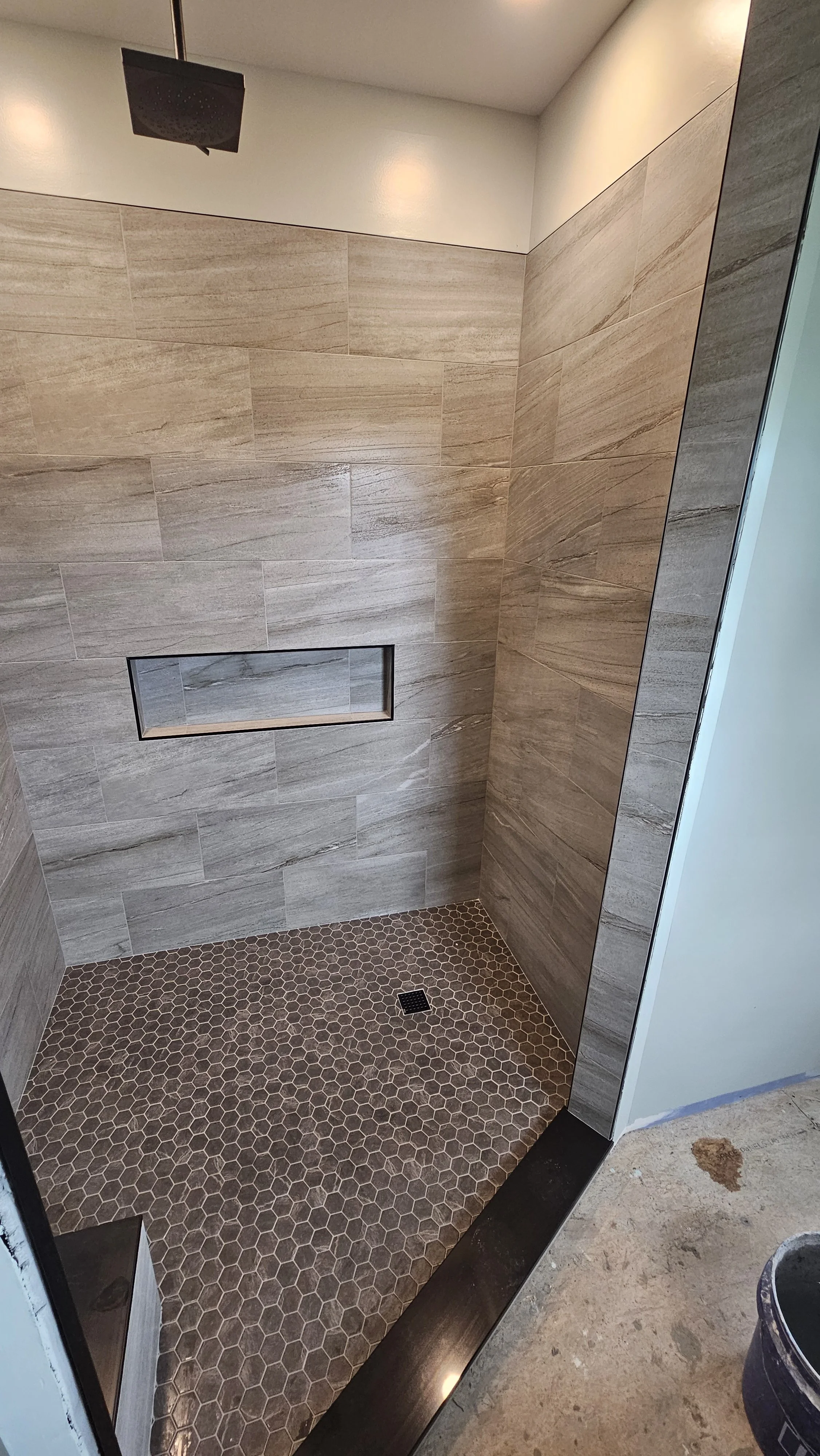 A modern shower with large beige tile walls, a built-in rectangular niche, hexagonal brown floor tiles, a black showerhead, and a floor drain.