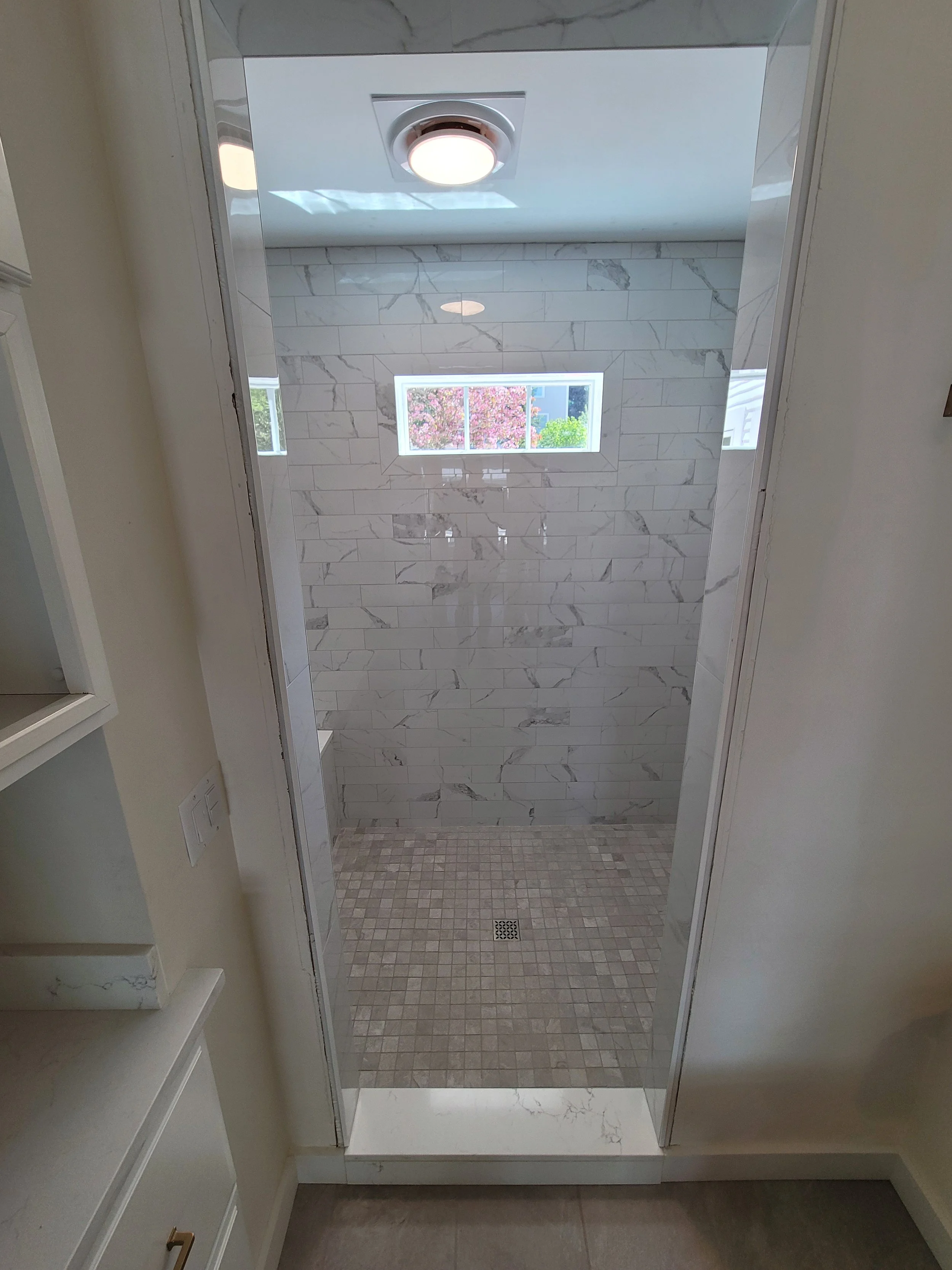 View of a modern walk-in shower with marble walls, a small window showing trees outside, and a tiled floor with a drain.