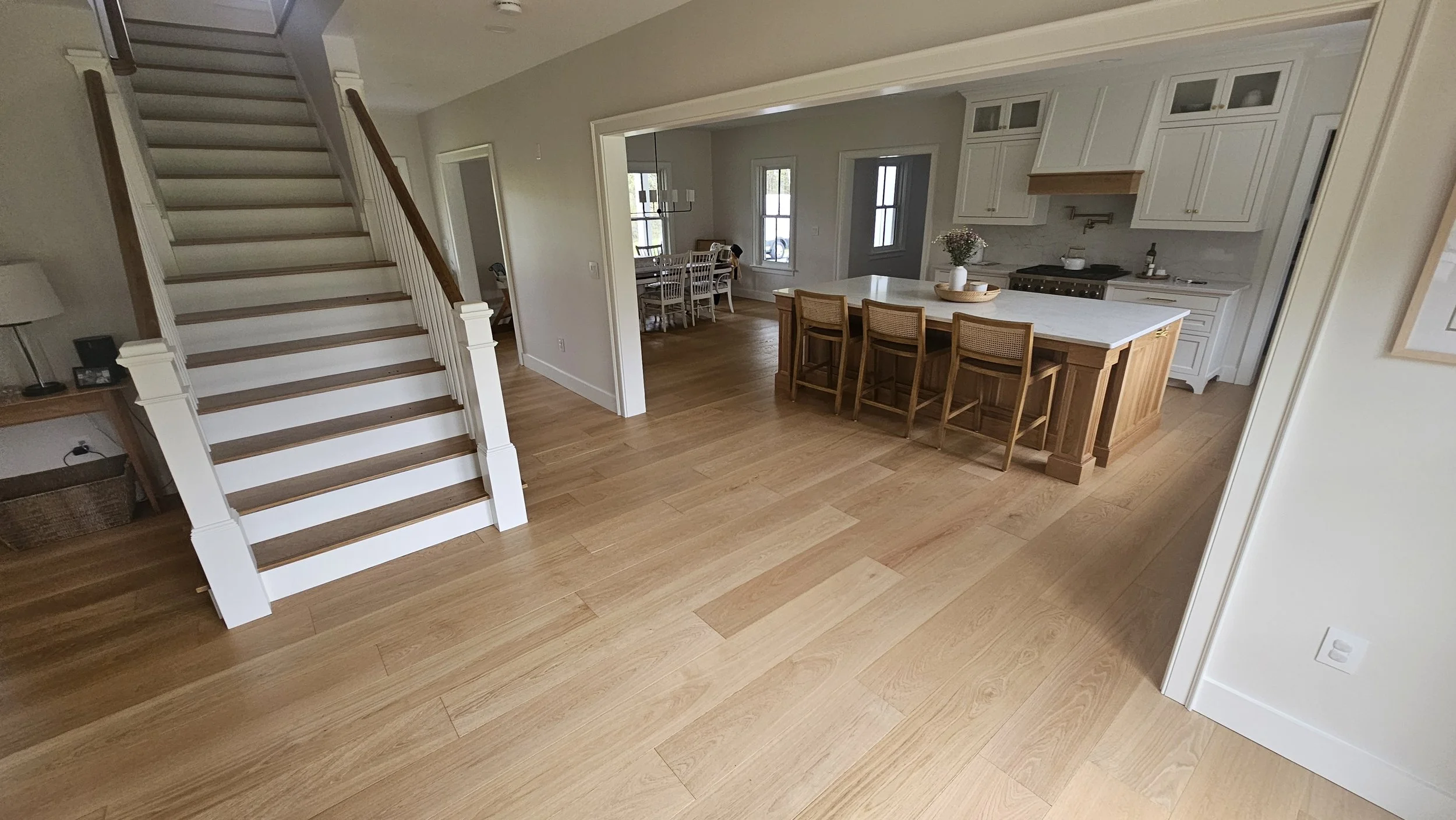 Open-concept kitchen and dining area with a staircase, wooden floors, white kitchen cabinets, an island with barstools, and a dining table with chairs.