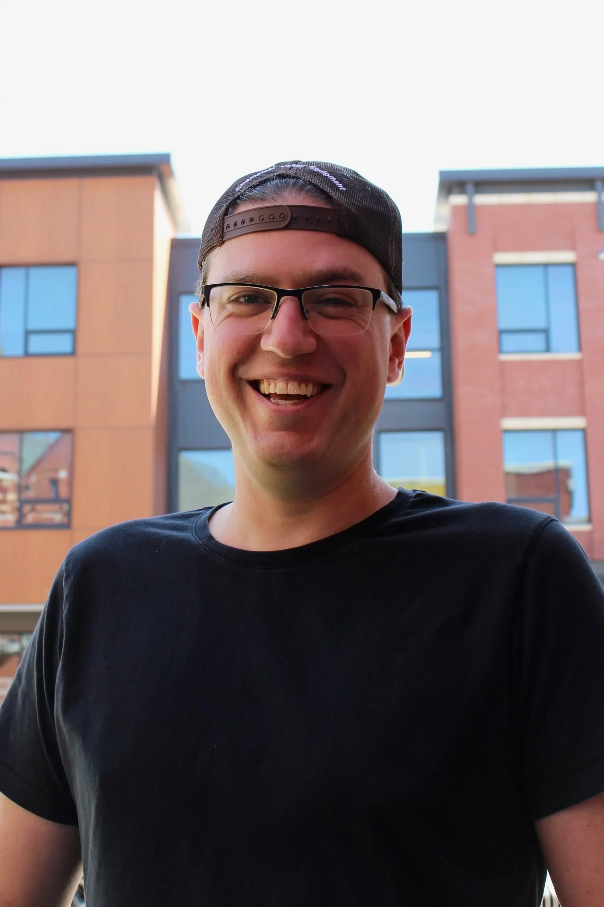 A young man smiling outdoors in front of a building with large windows and brick facades, wearing a black T-shirt, glasses, and a backward cap.