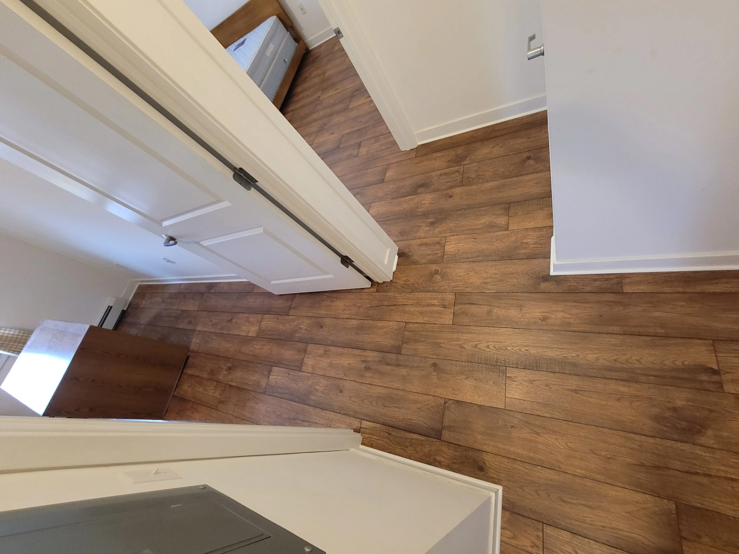Interior view of a room with wooden flooring, a white wall, and an open white door revealing a laundry room with a stacked washer and dryer.