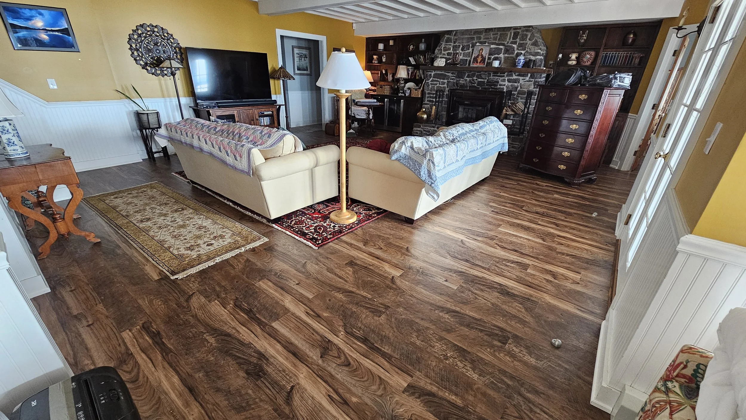 Living room with hardwood flooring, yellow painted walls, a stone fireplace with a wooden mantel, a large flat-screen TV on a wooden stand, two cream-colored sofas with blankets, a floor lamp, a decorative wall piece, and various paintings and booksh