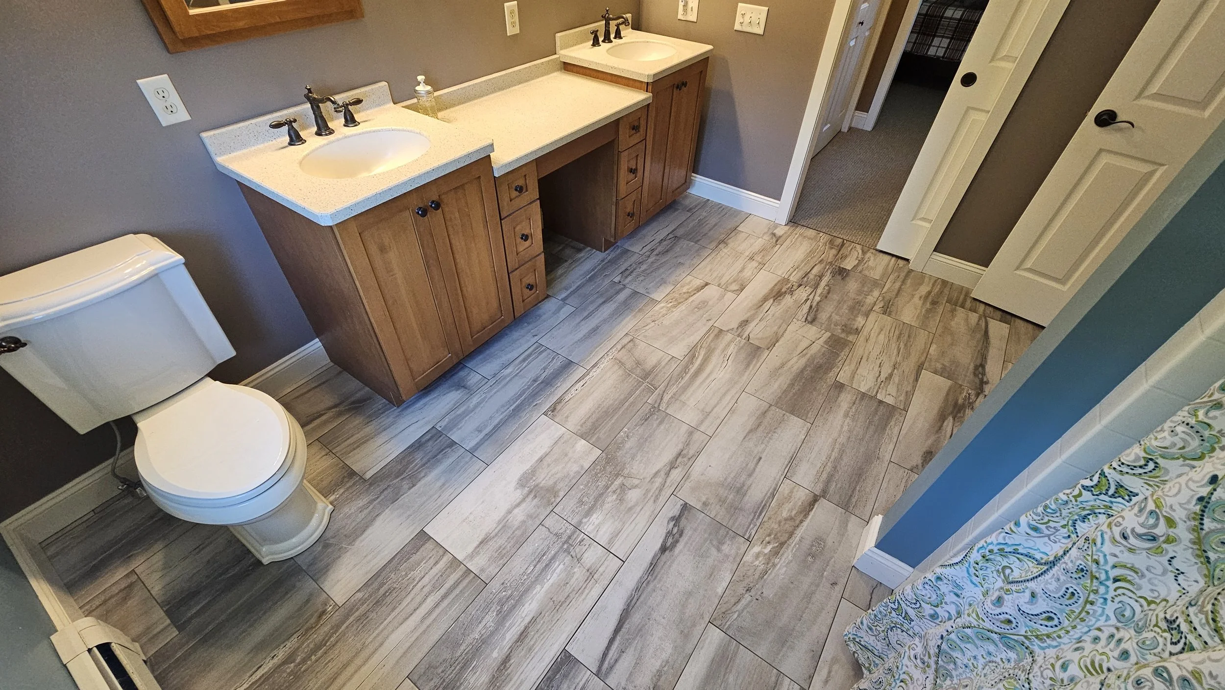 Bathroom with double vanity sinks, wooden cabinets, and a toilet, with gray walls and a patterned curtain.