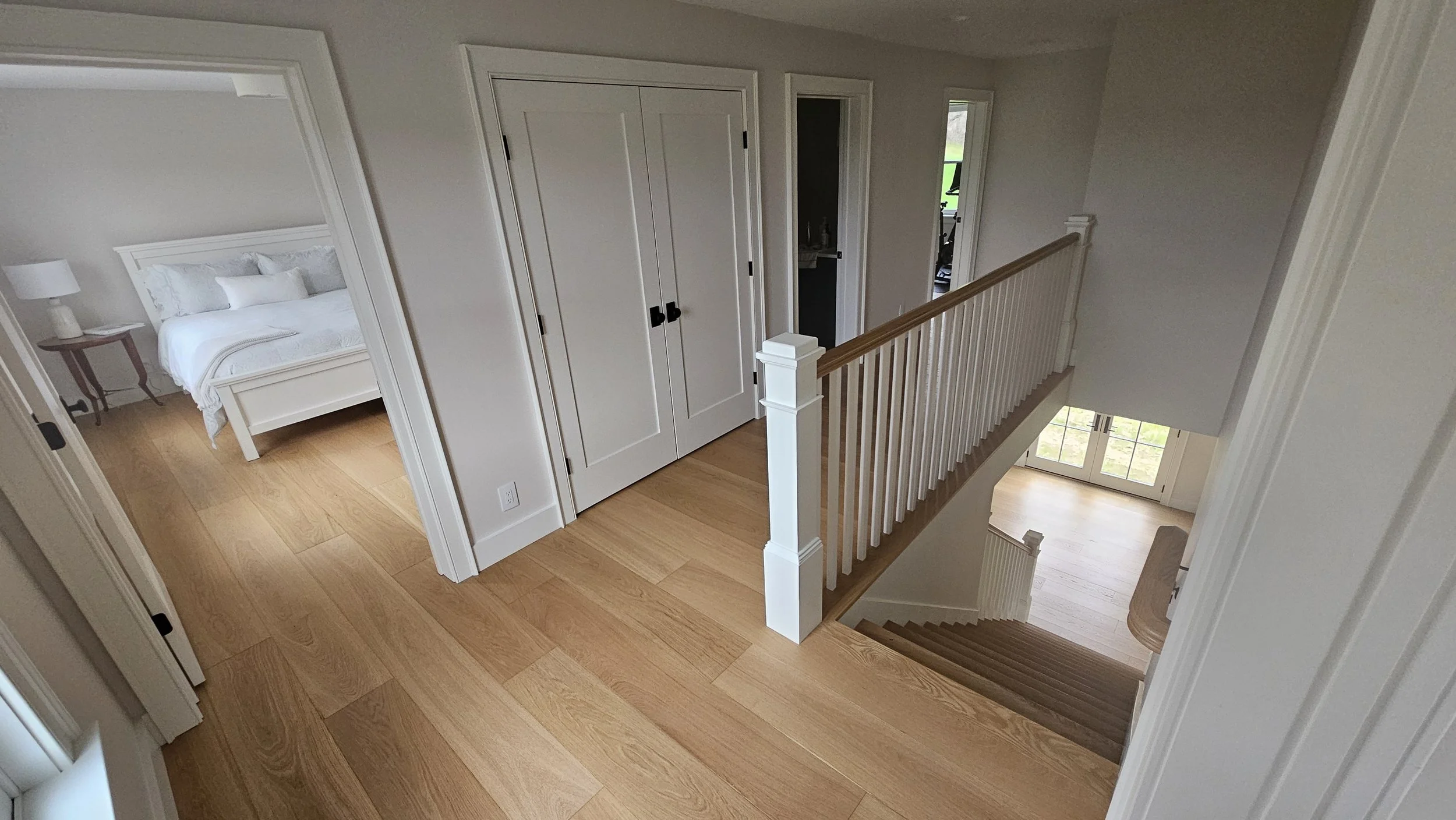 Interior view of a modern house with light-colored wooden floors, white walls, staircase, and multiple rooms including a bedroom with white bedding and a dark wood side table. Multiple doors lead to other rooms, and there is natural light coming from