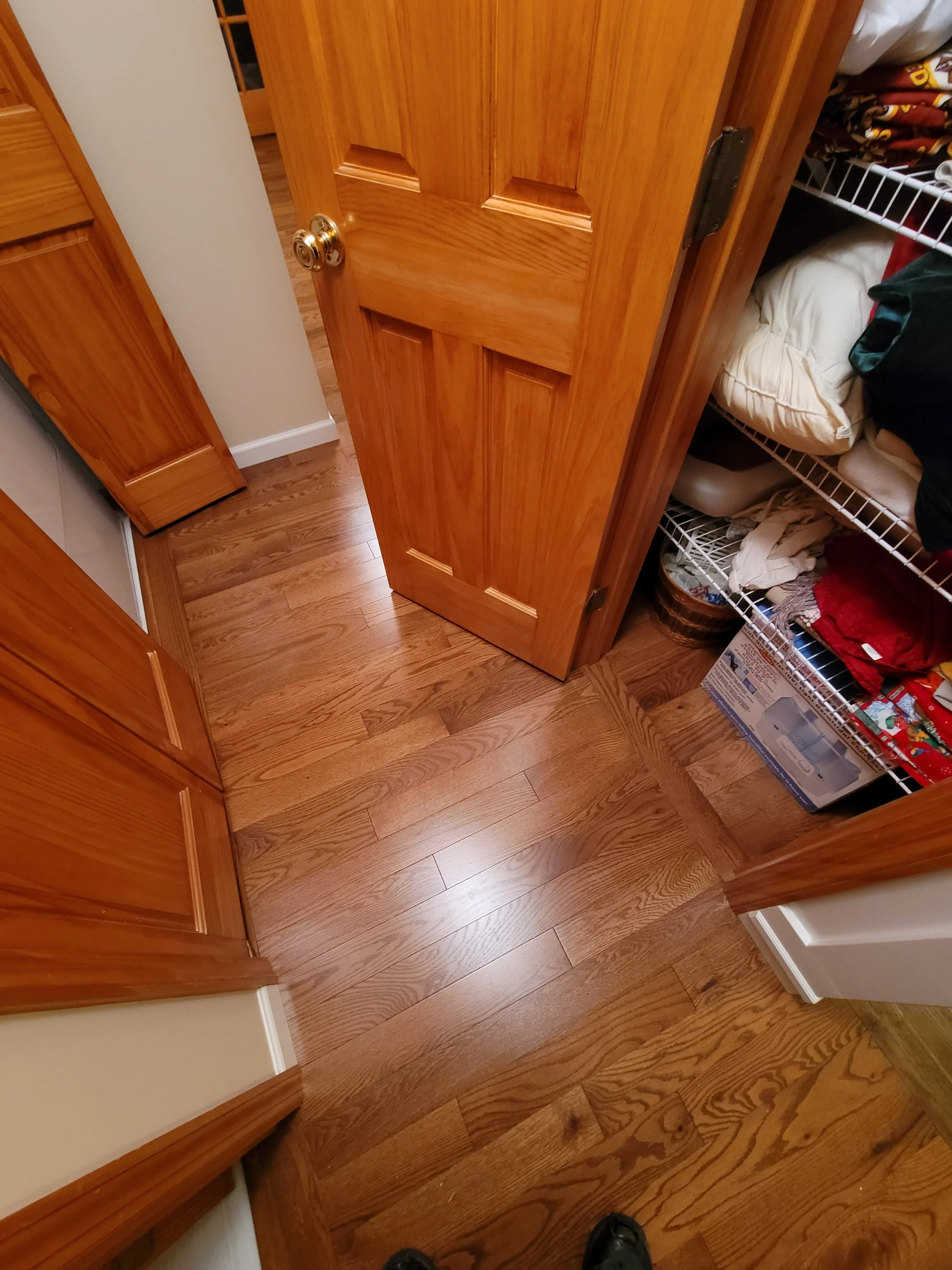 A wooden pantry door slightly ajar with a wire shelf and various items inside, including blankets and boxes, in a kitchen with hardwood flooring.