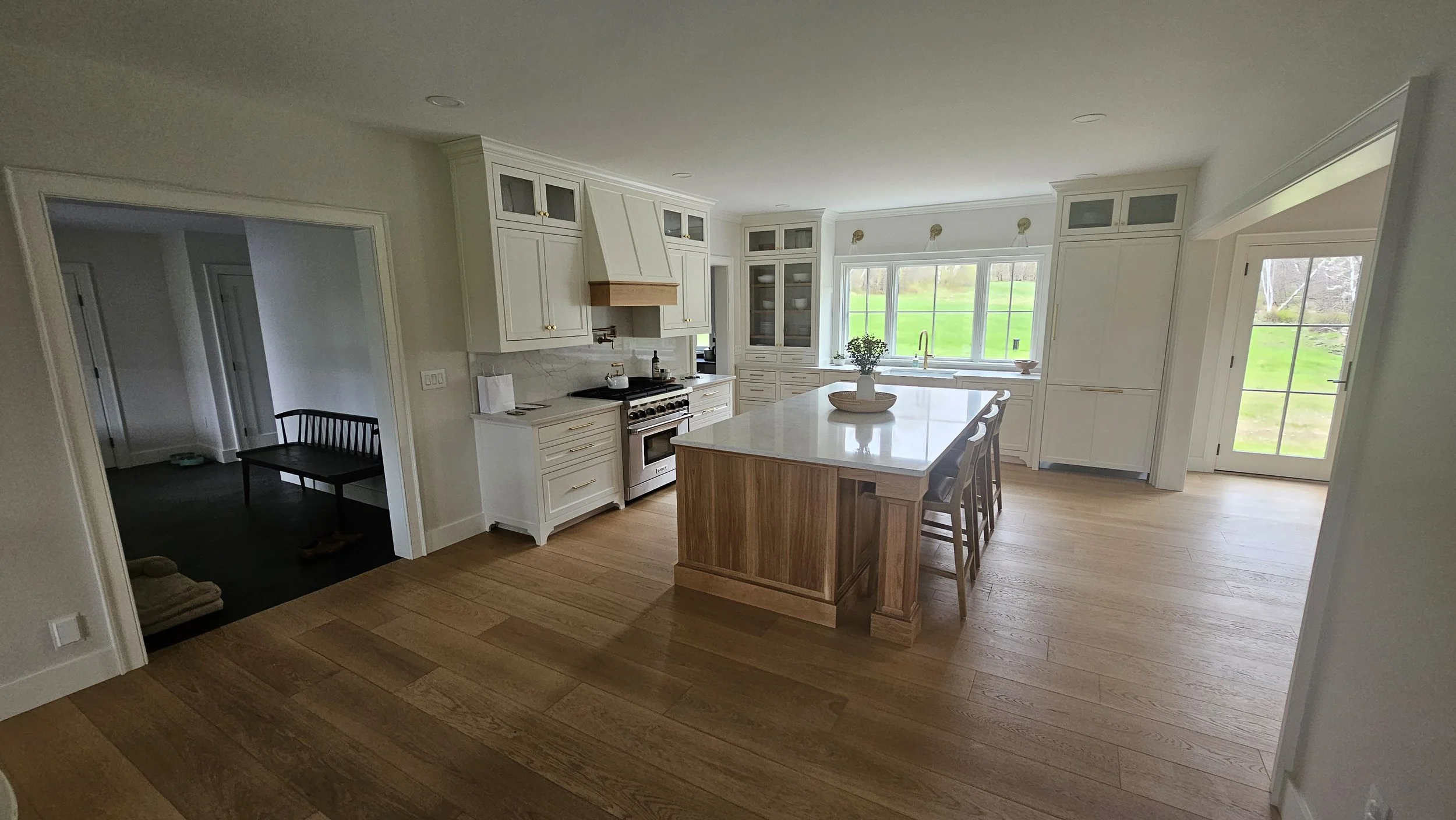 A spacious kitchen with white cabinetry, marble backsplash, a central island with a wooden base, and large windows overlooking a green yard.