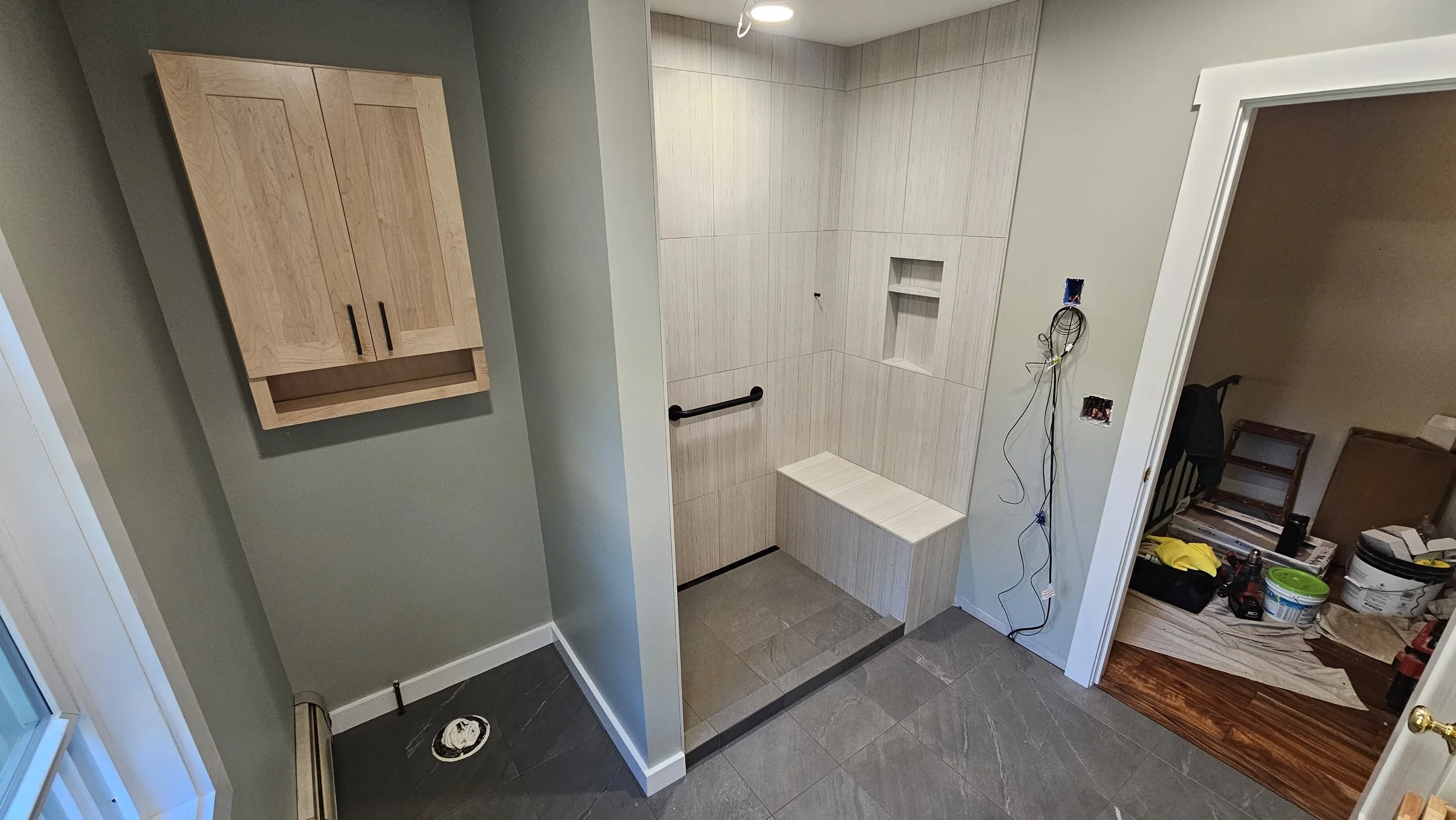 Bathroom under renovation with beige tile shower area, built-in shelf, and black grab bar, adjacent to a light green wall-mounted cabinet, with construction tools and supplies visible.