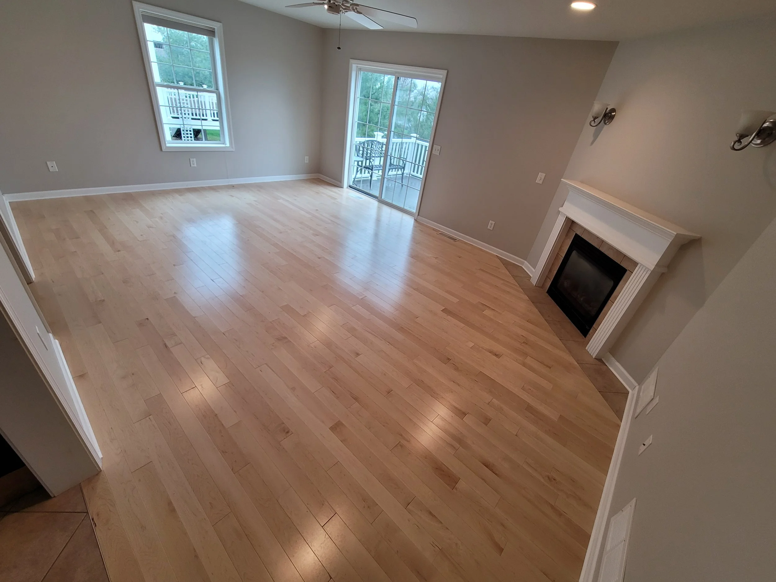 Empty living room with wooden floors, two windows, a sliding glass door leading to a small balcony, a fireplace, and beige walls.