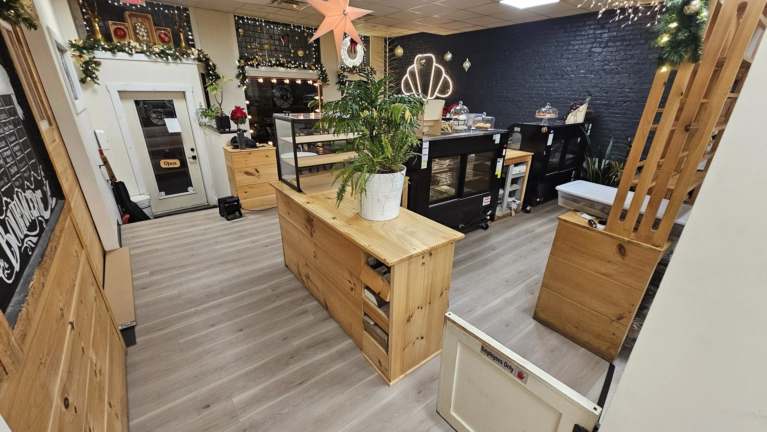 Interior of a cozy establishment decorated for Christmas, featuring a black brick wall, garlands, ornaments, and a star-shaped light fixture. There is a wooden counter with a large potted plant, display cases with baked goods, and a chalkboard menu o