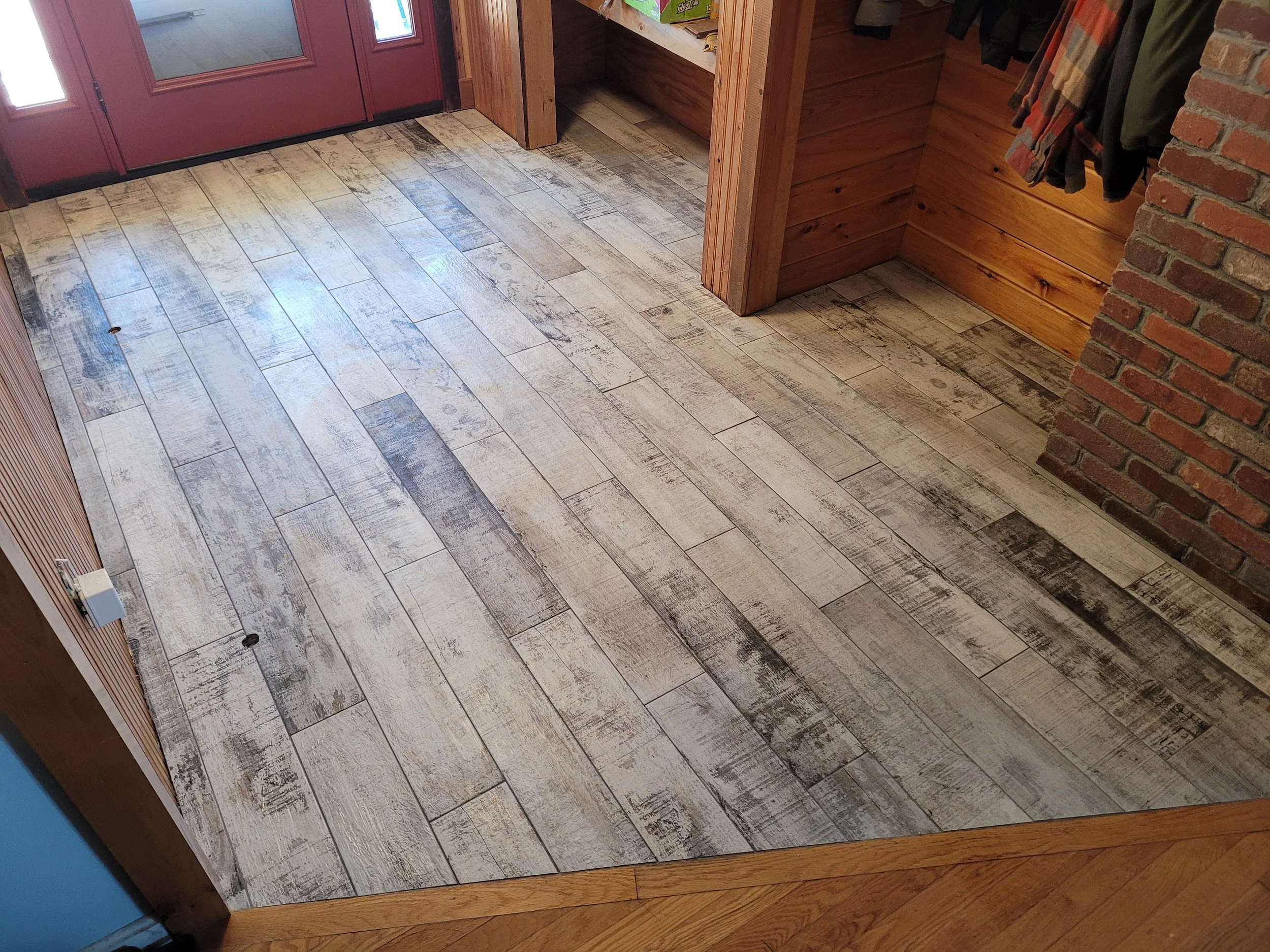 Wooden flooring with a six-inch plank distressed finish and a corner of a brick wall on the right side, with a wooden trim at the base of the wall and a red door with glass panels.