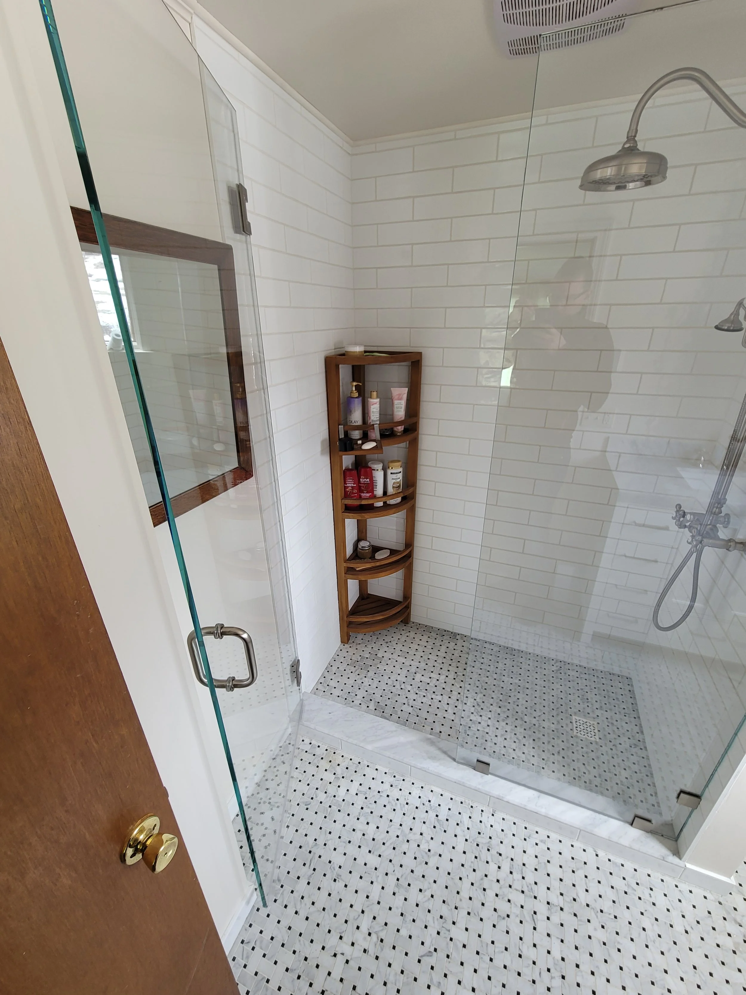 Bathroom with glass shower enclosure, white subway tile walls, small black and white mosaic floor tiles, wooden corner shelf with toiletries, shower head, faucet, and a mirror.
