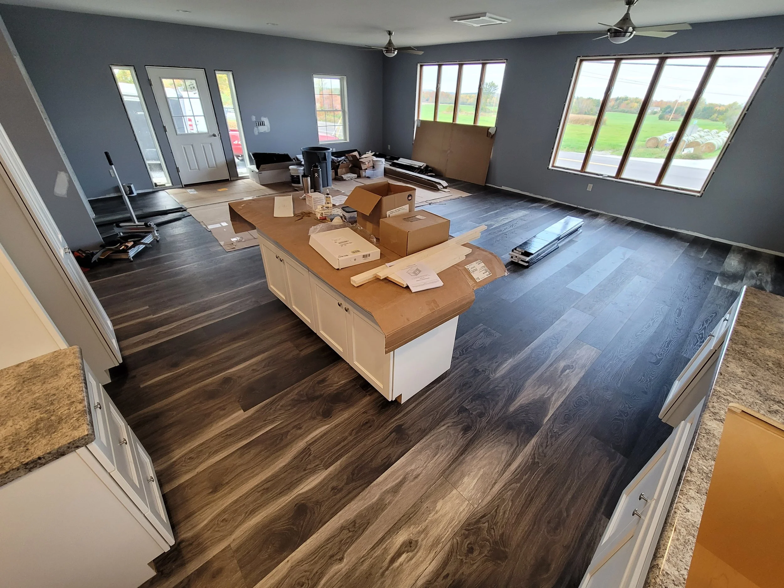 Living room under construction with tools, boxes, and materials scattered on the floor. Large windows with a view of a green field and hay bales outside.