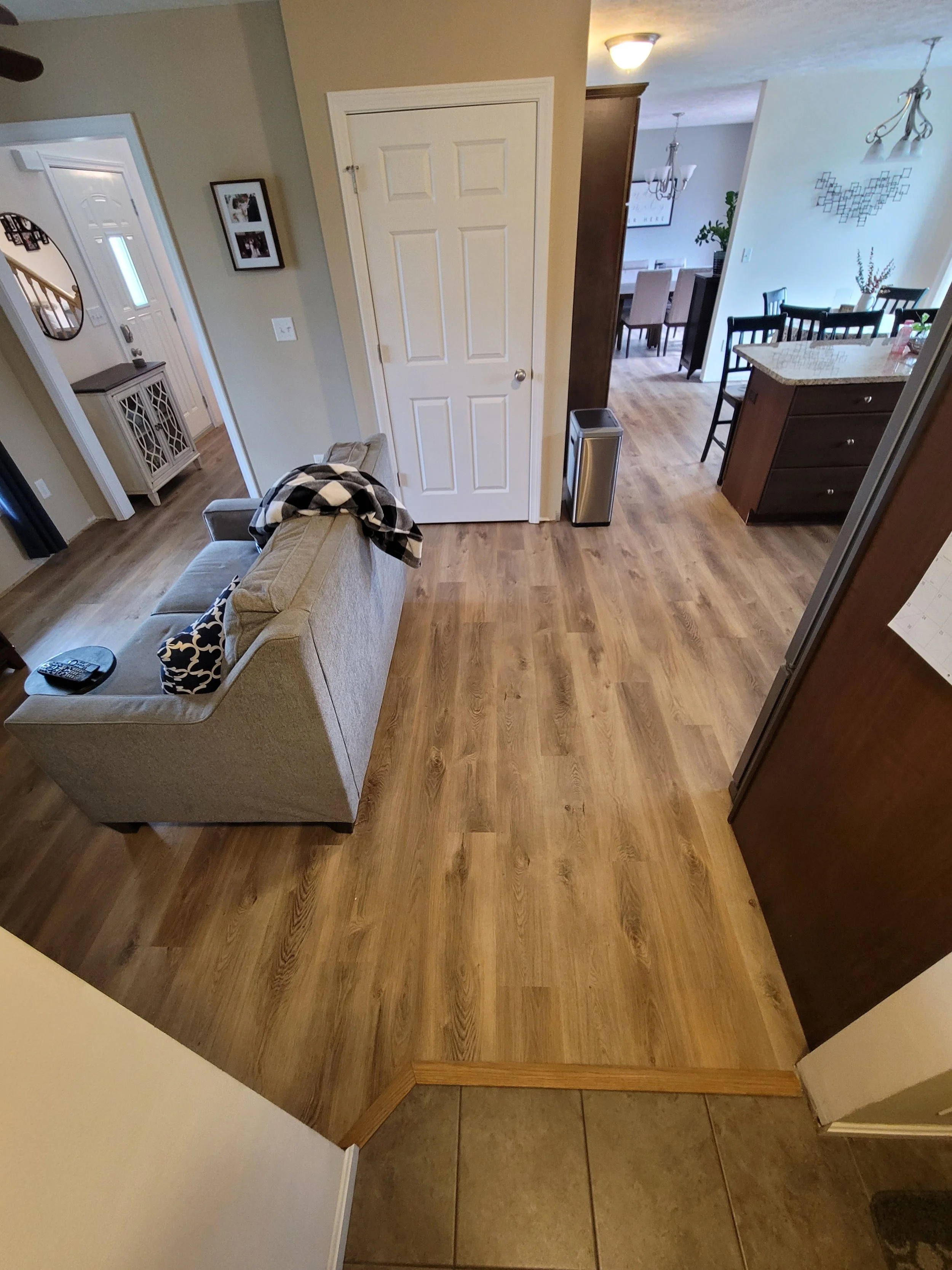 View of a living room with a beige couch, a small table, a wall with picture frames, and a wooden floor transitioning to a tiled entryway.
