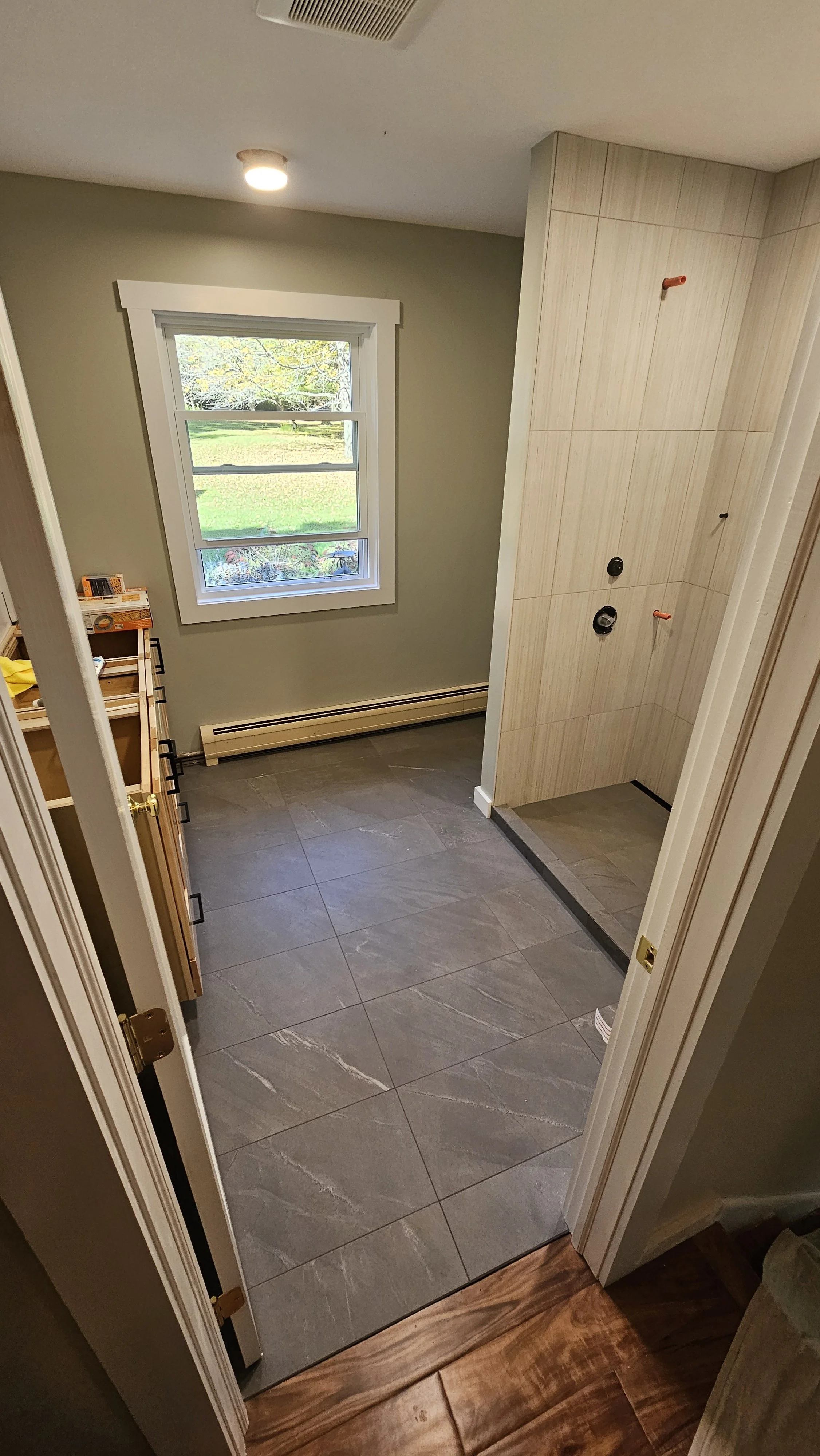 Bathroom with gray tiled floor, a window overlooking a backyard with trees, and an unfinished shower area with walls and plumbing fixtures, seen through a door.