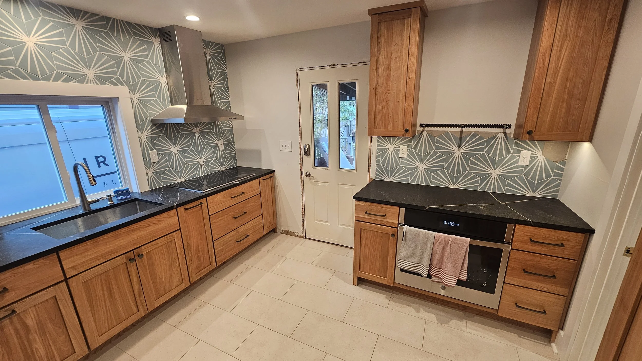 Kitchen with wooden cabinets, black countertops, patterned backsplash, stainless steel range hood, window over sink, and a stainless steel oven.