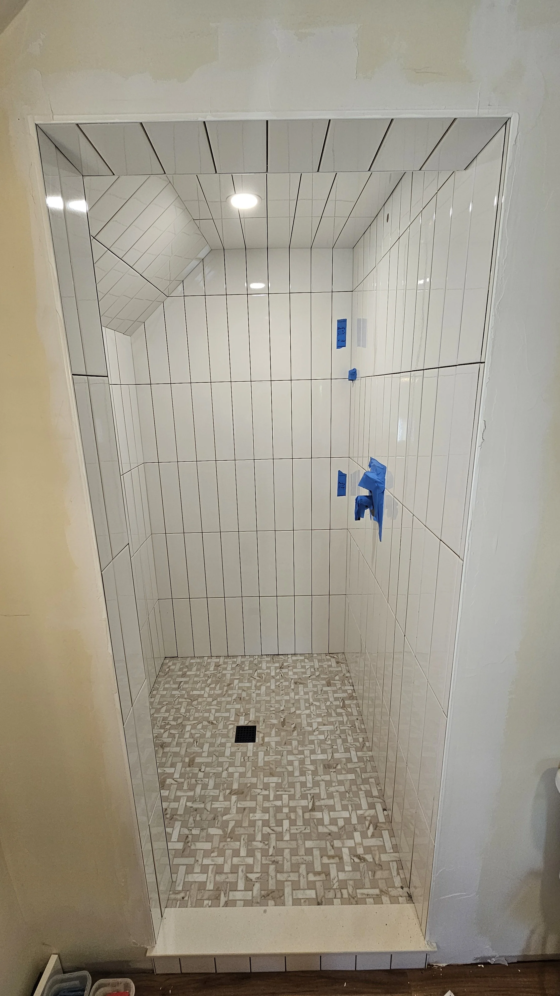 Interior of a shower with white tile walls, a small patterned tile floor, and a recessed ceiling light, missing fixtures, with blue painter's tape on the wall.