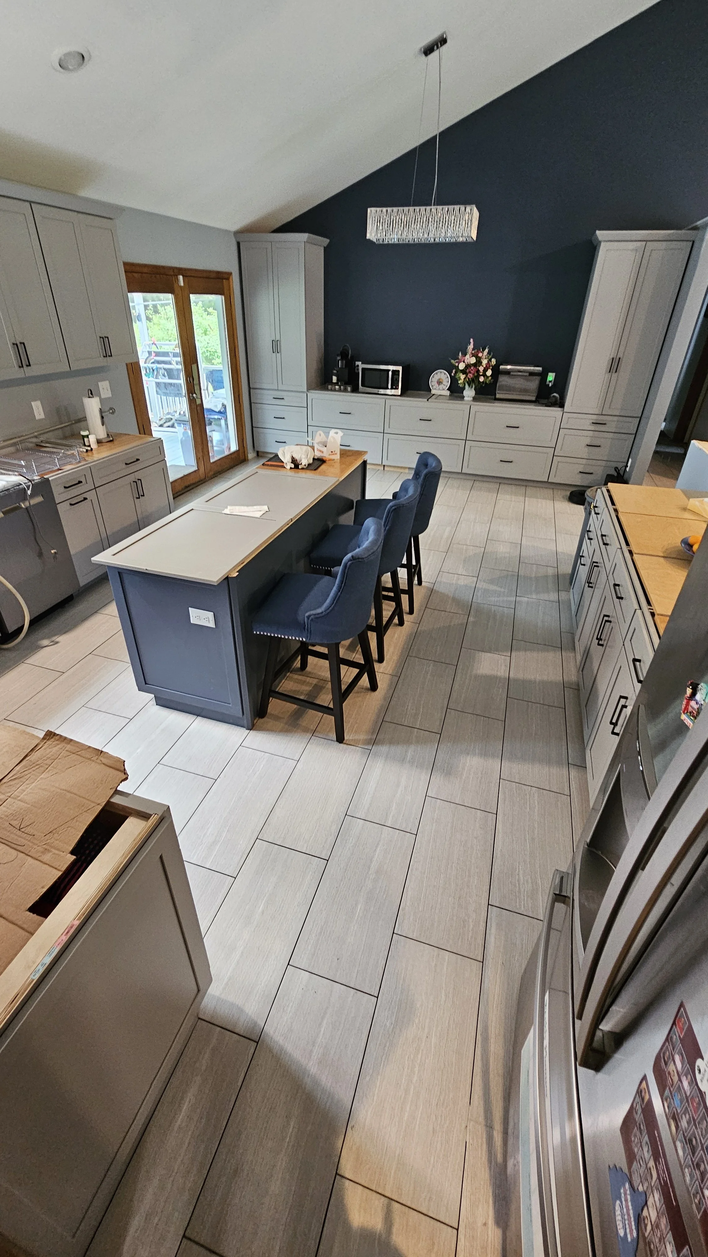 Modern kitchen with light gray cabinets, a large kitchen island with three blue upholstered barstools, a dark accent wall, and a chandelier overhead. Features include a microwave, flowers, and sliding glass doors leading outside.