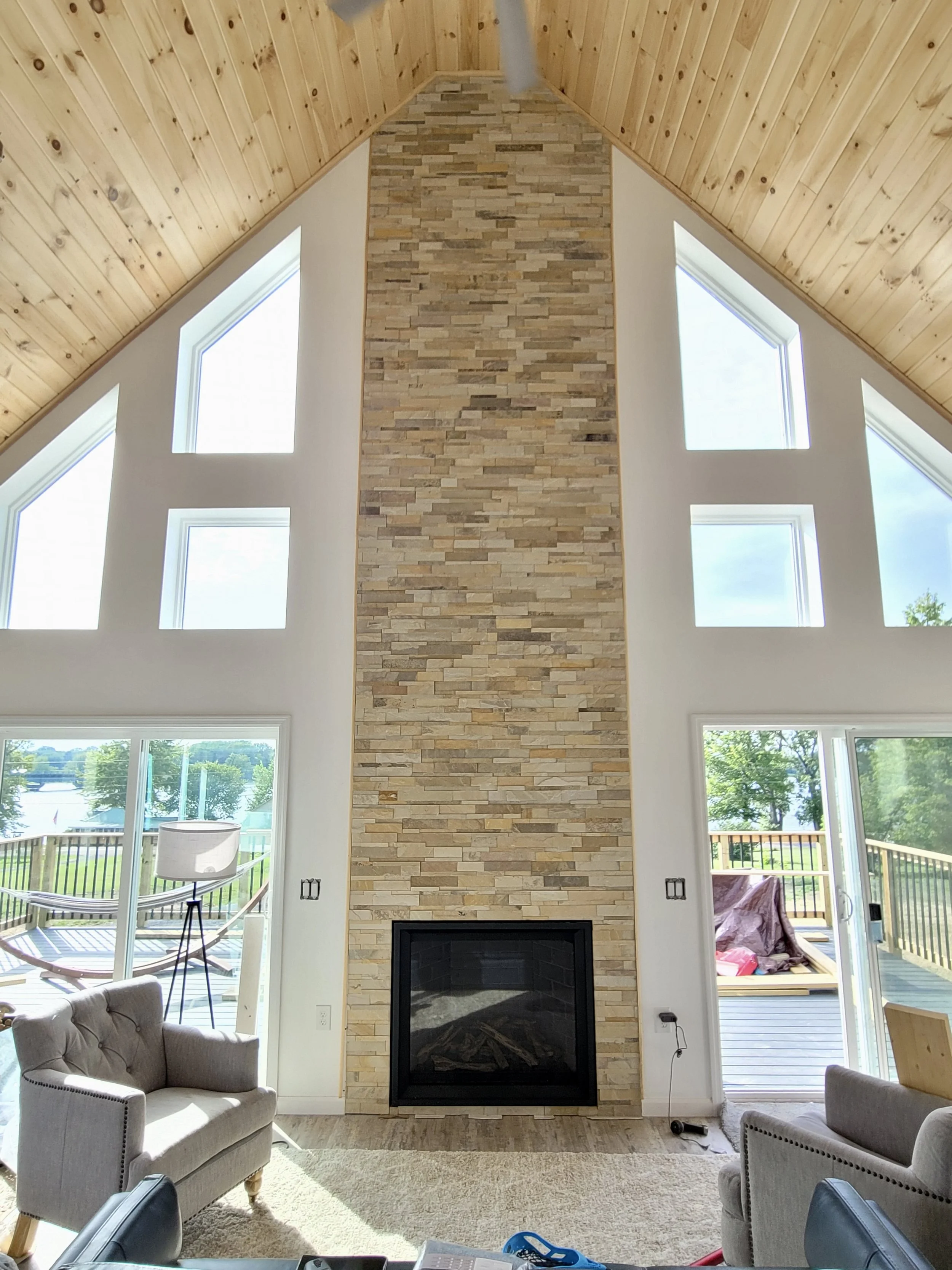 Living room with a wood-paneled vaulted ceiling, two large windows on each side of a stone fireplace, and an outdoor deck visible through glass doors.