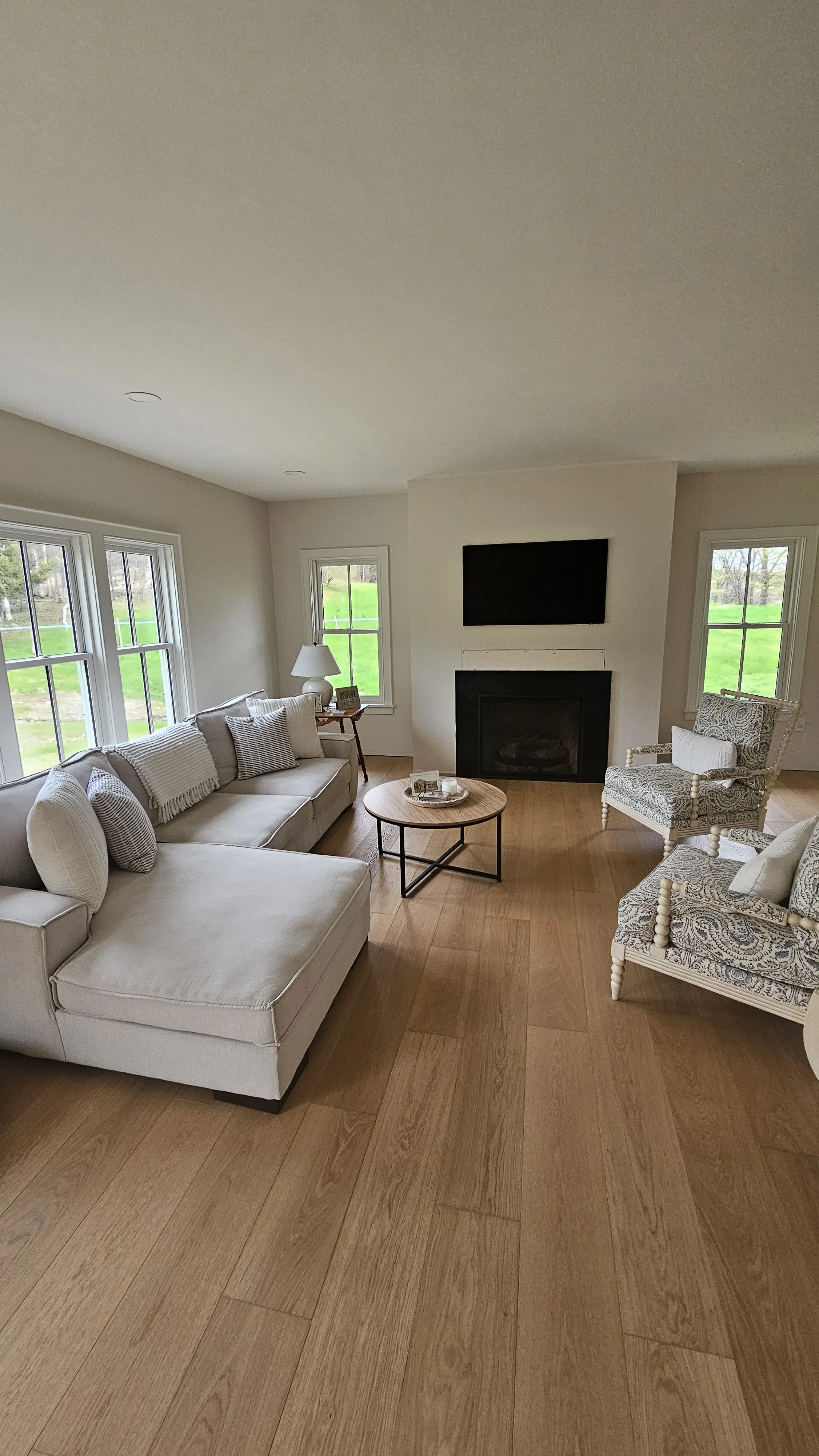 Living room with cream sofa, patterned armchairs, wood flooring, fireplace, mounted TV, windows with greenery outside, and a small round coffee table.