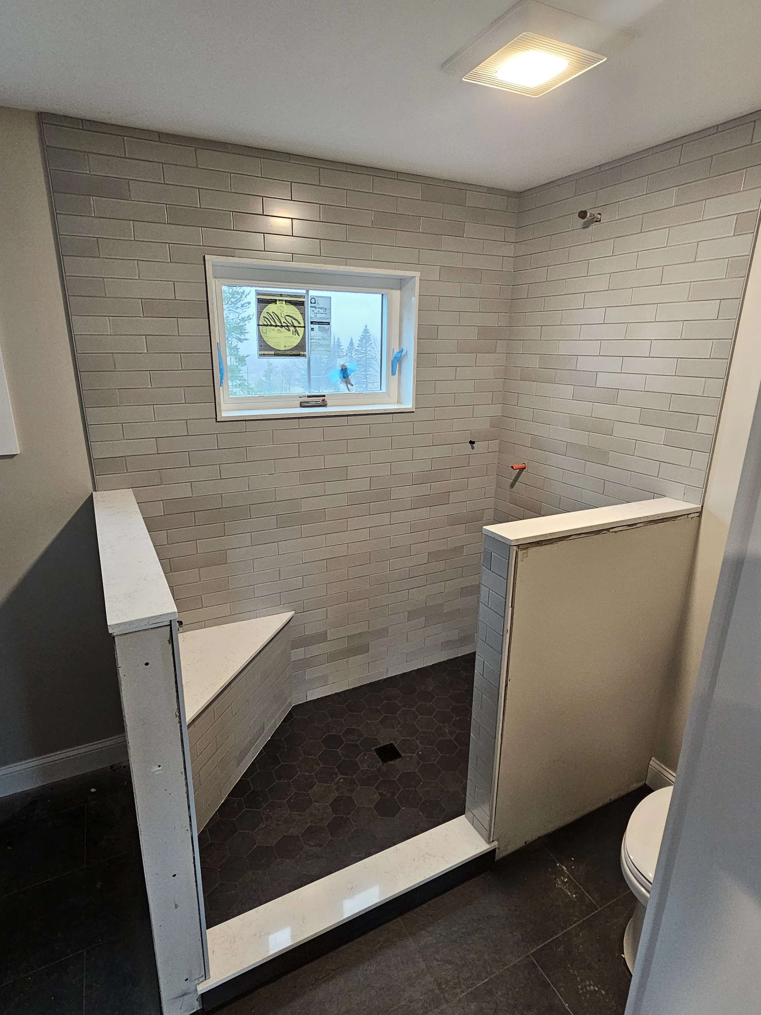 A bathroom with striped beige tile walls, a small window, and a black hexagonal tile floor. There is a white toilet in the corner and partially installed partitions or shelves.